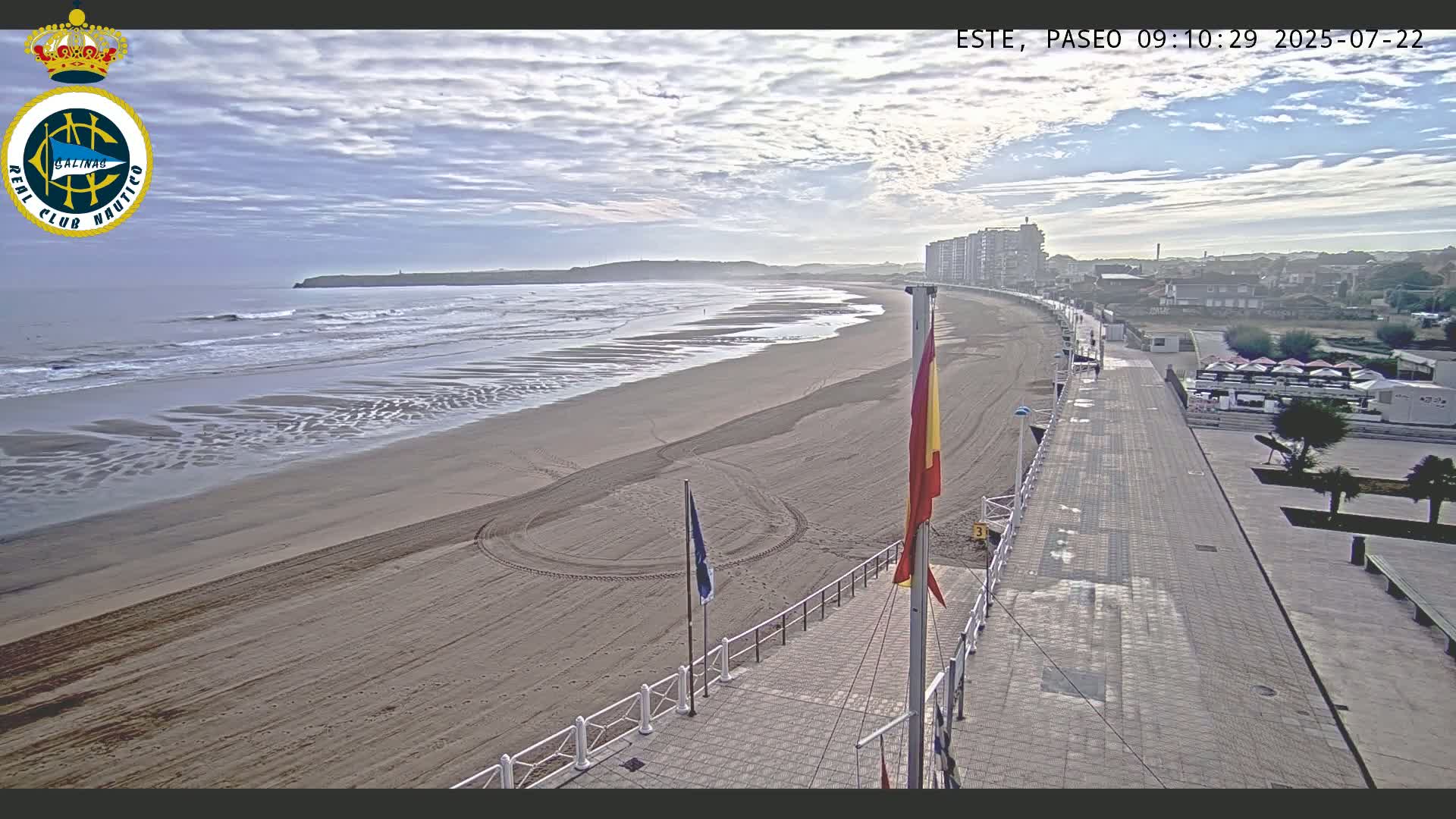 A sandy beach with gentle waves under a partly cloudy sky is viewed from a boardwalk overlooking the ocean and a town in the distance.
