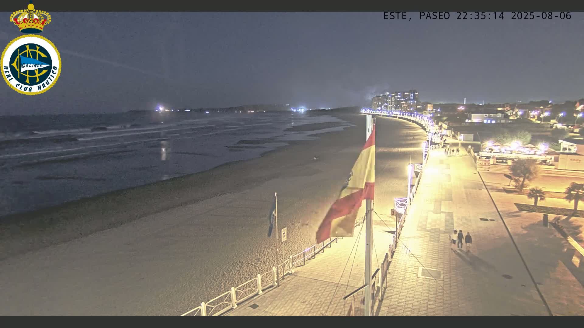 A nighttime beach scene shows a boardwalk with people walking along it,  illuminated buildings in the background, and gentle waves lapping the shore under a dark sky.