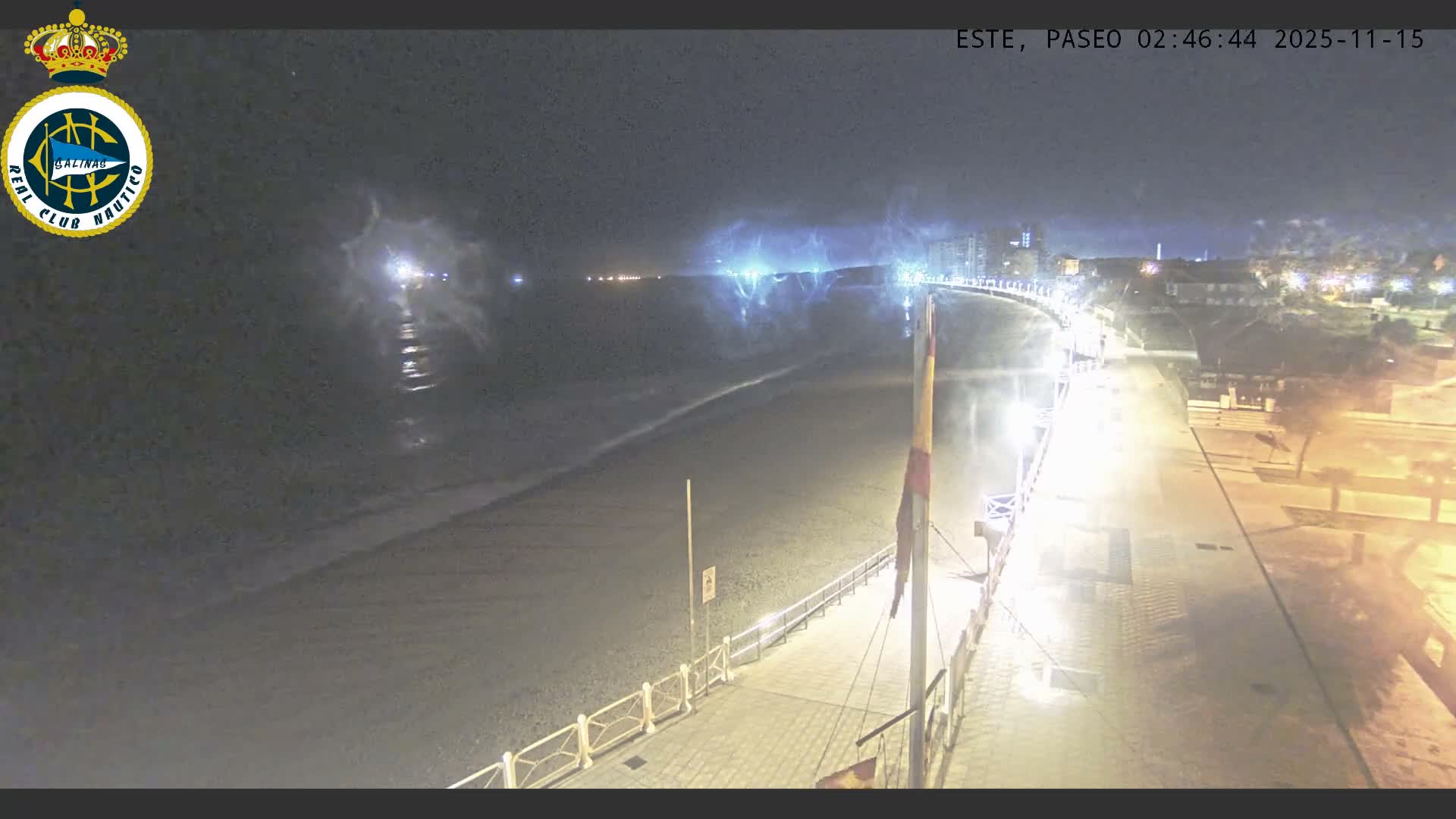 A nighttime view reveals a wide, sandy beach alongside an illuminated promenade, with distant city lights and a significant atmospheric haze around bright blue lights over the water.