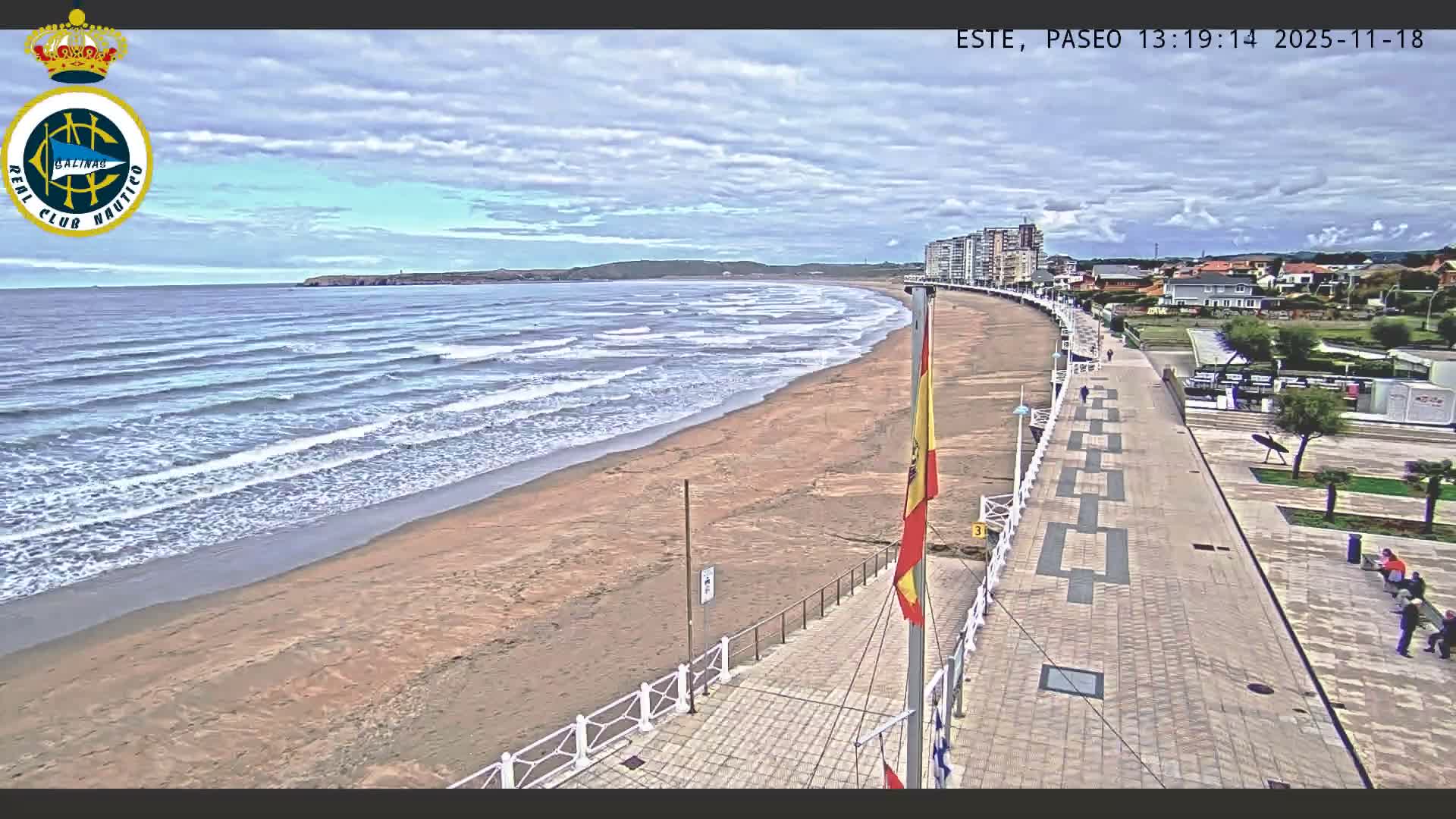 A nighttime view reveals a wide, sandy beach alongside an illuminated promenade, with distant city lights and a significant atmospheric haze around bright blue lights over the water.