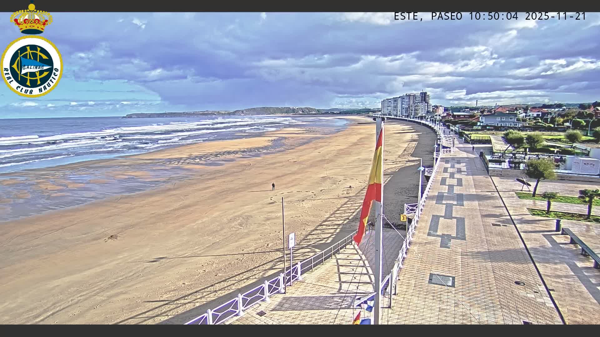 A wide panoramic view captures a sandy beach with breaking waves, a lone person walking on the shore, and a long patterned promenade bordered by a railing, all beneath a partly cloudy sky with distant coastal hills and buildings.