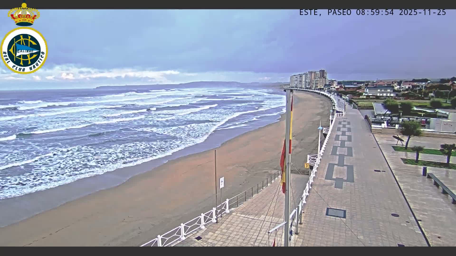 A wide sandy beach with breaking waves is bordered by a paved boardwalk and a town with buildings, all under an overcast sky with distant hills.