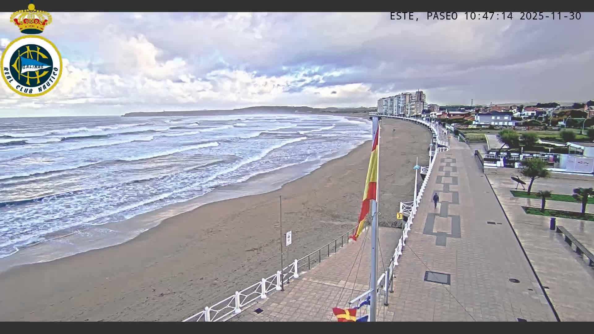 A wide sandy beach with breaking waves stretches along a coastline featuring a promenade, buildings, and a flying Spanish flag, all under a heavily overcast sky.