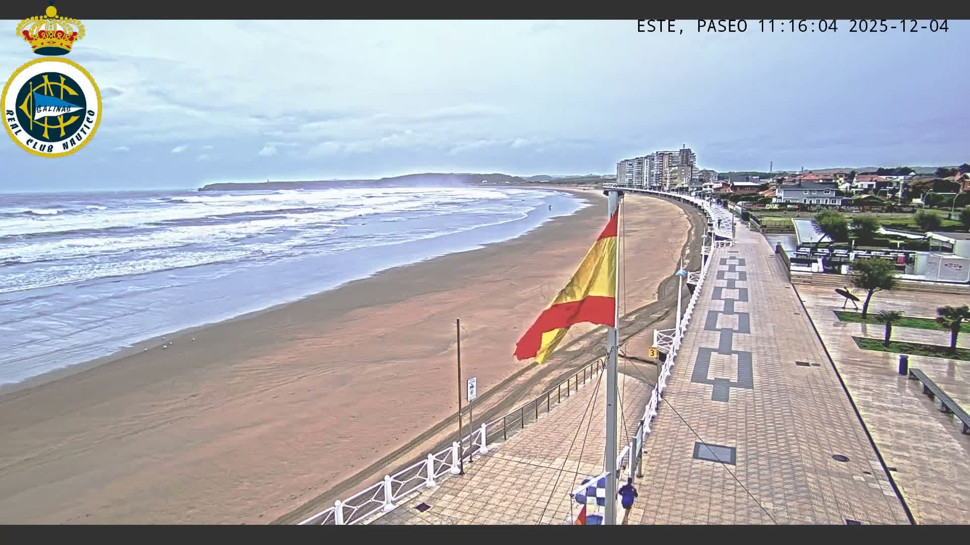A wide sandy beach with breaking waves extends along a patterned promenade under an overcast sky, featuring a flying Spanish flag and coastal buildings in the distance.