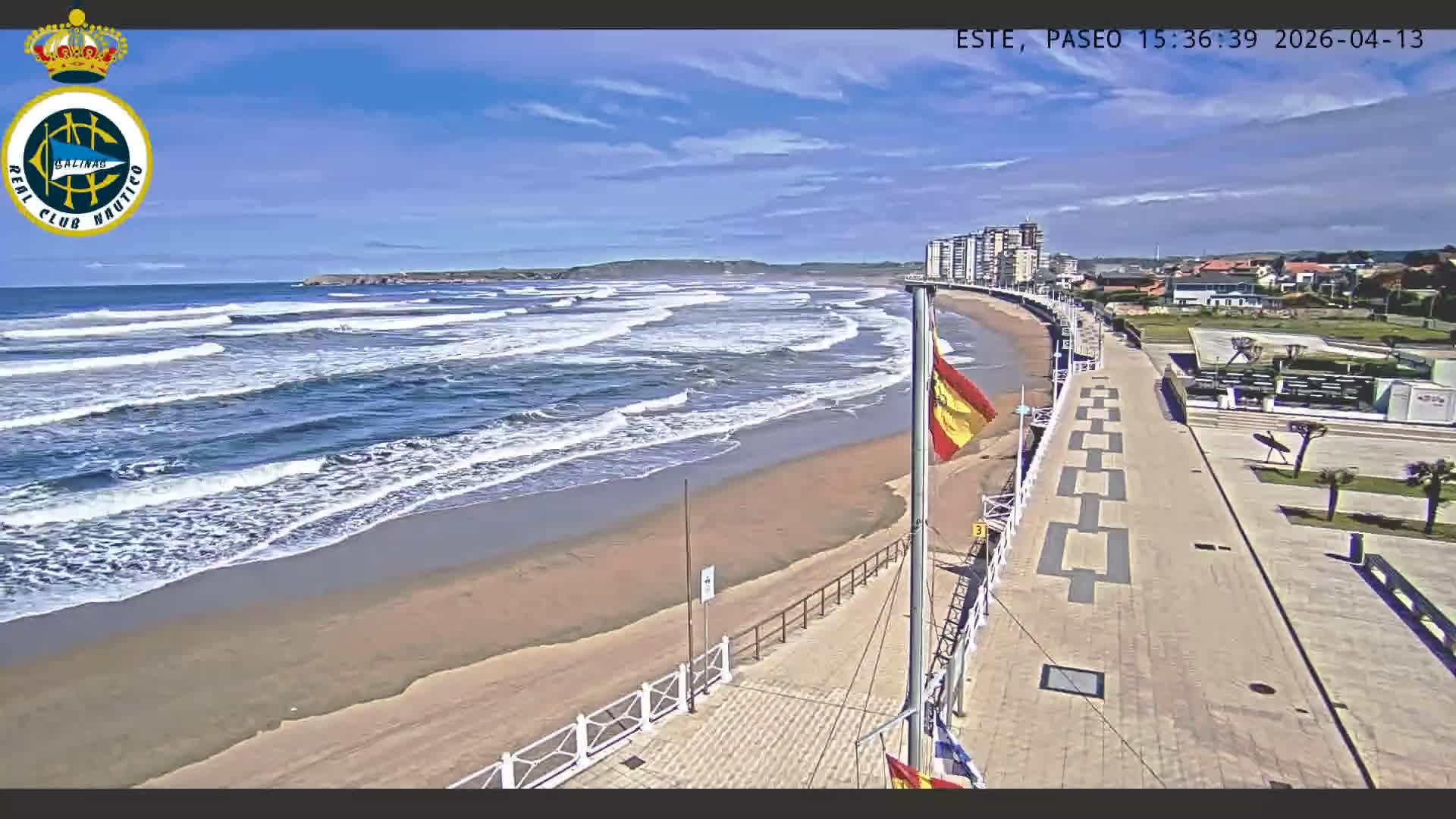 A wide sandy beach with breaking waves extends along a patterned promenade under an overcast sky, featuring a flying Spanish flag and coastal buildings in the distance.