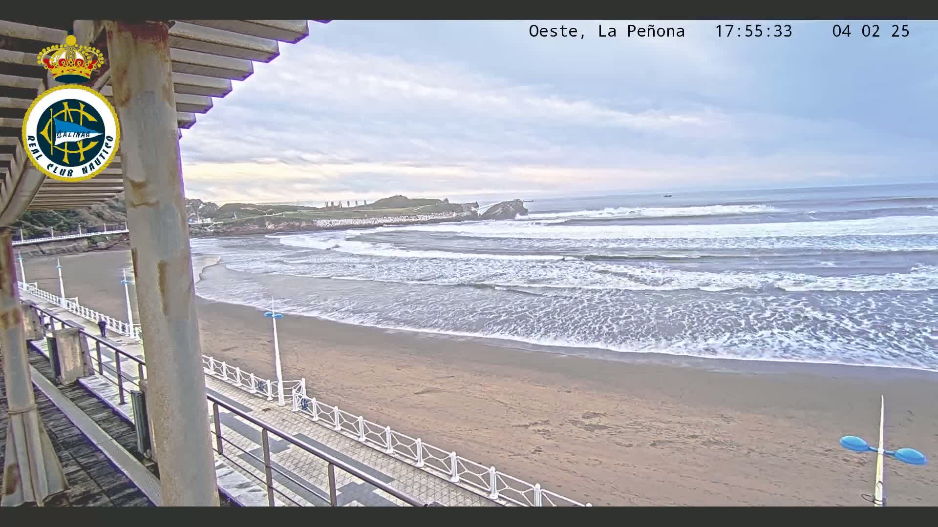 A sandy beach with gentle waves under a cloudy sky is viewed from an elevated position, showing a boardwalk and a distant rocky coastline.