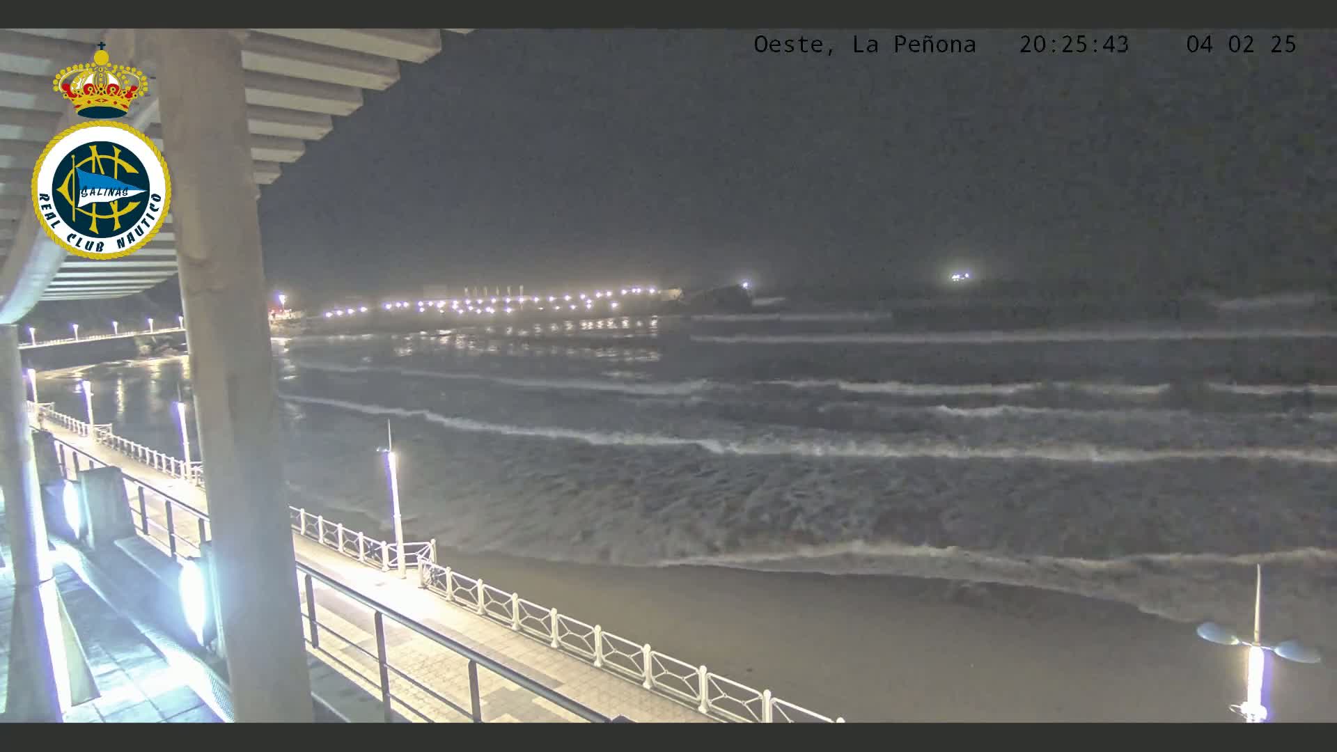 A nighttime view of a beach with gently rolling waves, lit by lights along a boardwalk stretching from a building.