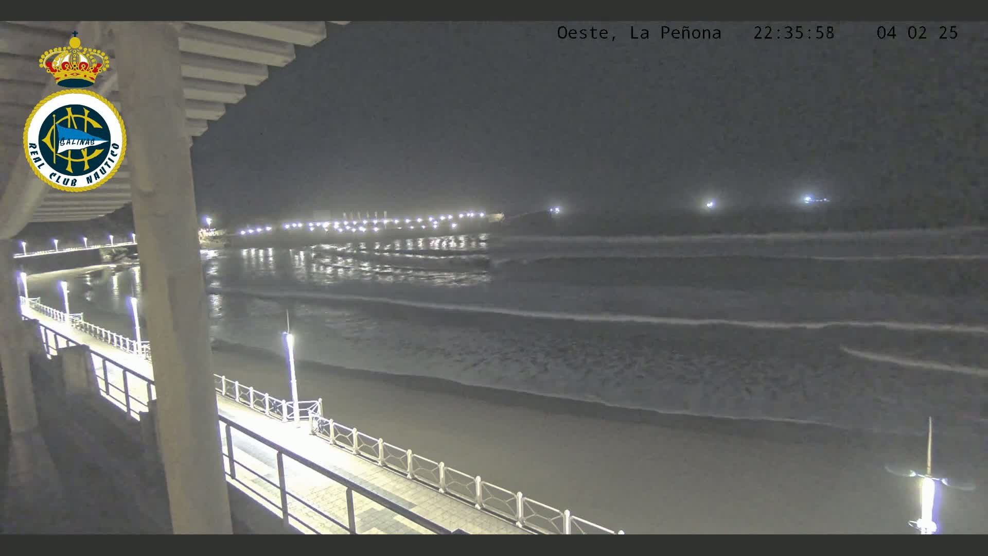 A nighttime view from a pier shows gently rolling waves on a dark beach under a clear sky, with lights illuminating the shoreline in the distance.