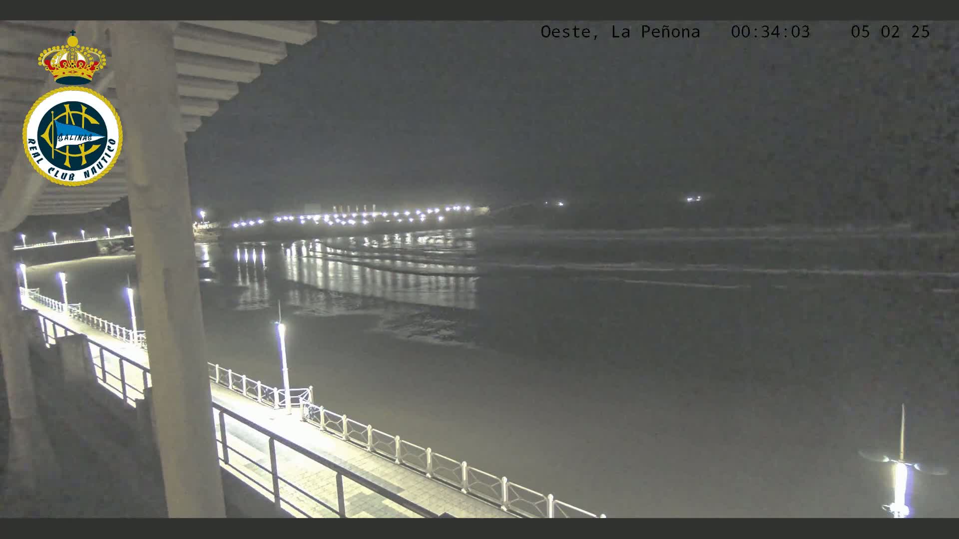 A nighttime view from a pier shows a calm, dark ocean reflecting the lights of a shoreline at a distance.
