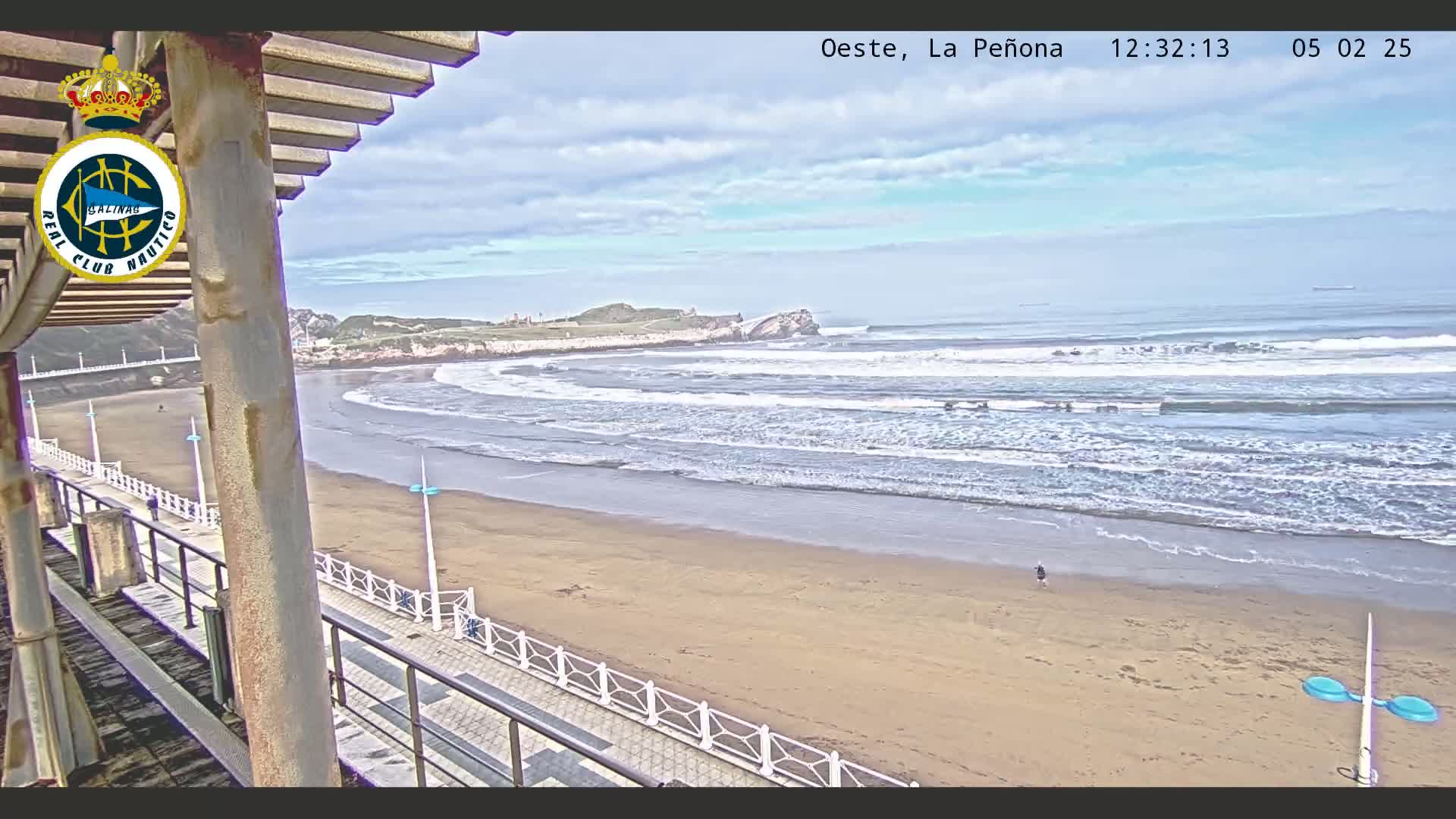 A sandy beach with gentle waves under a partly cloudy sky is viewed from an elevated position, overlooking a walkway and a distant rocky outcrop.