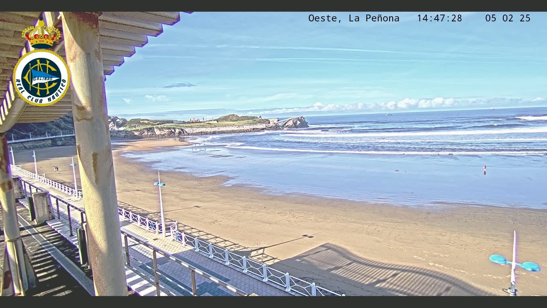 A mostly sunny day shows a sandy beach with gentle waves lapping the shore, viewed from an elevated position overlooking a boardwalk.