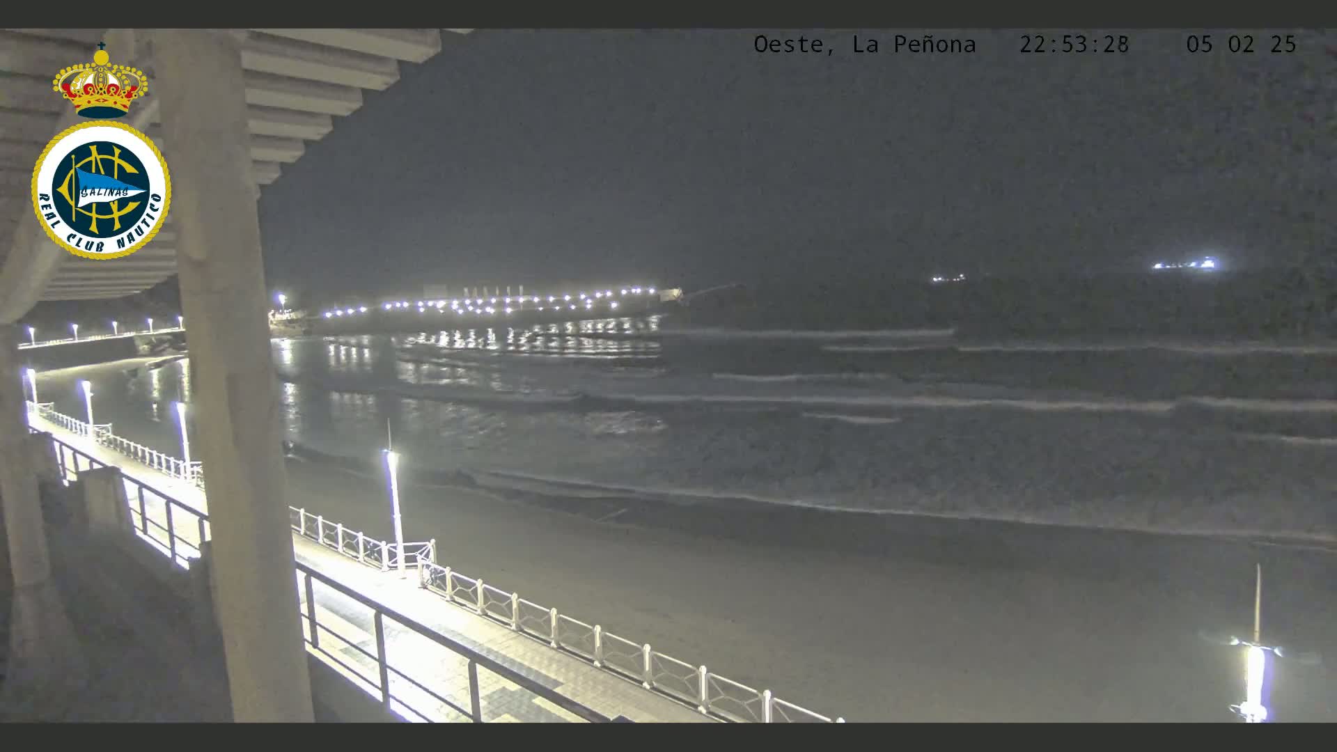 A nighttime view from an elevated vantage point shows a beach with gently rolling waves under a dark sky, and a lit walkway along a pier.