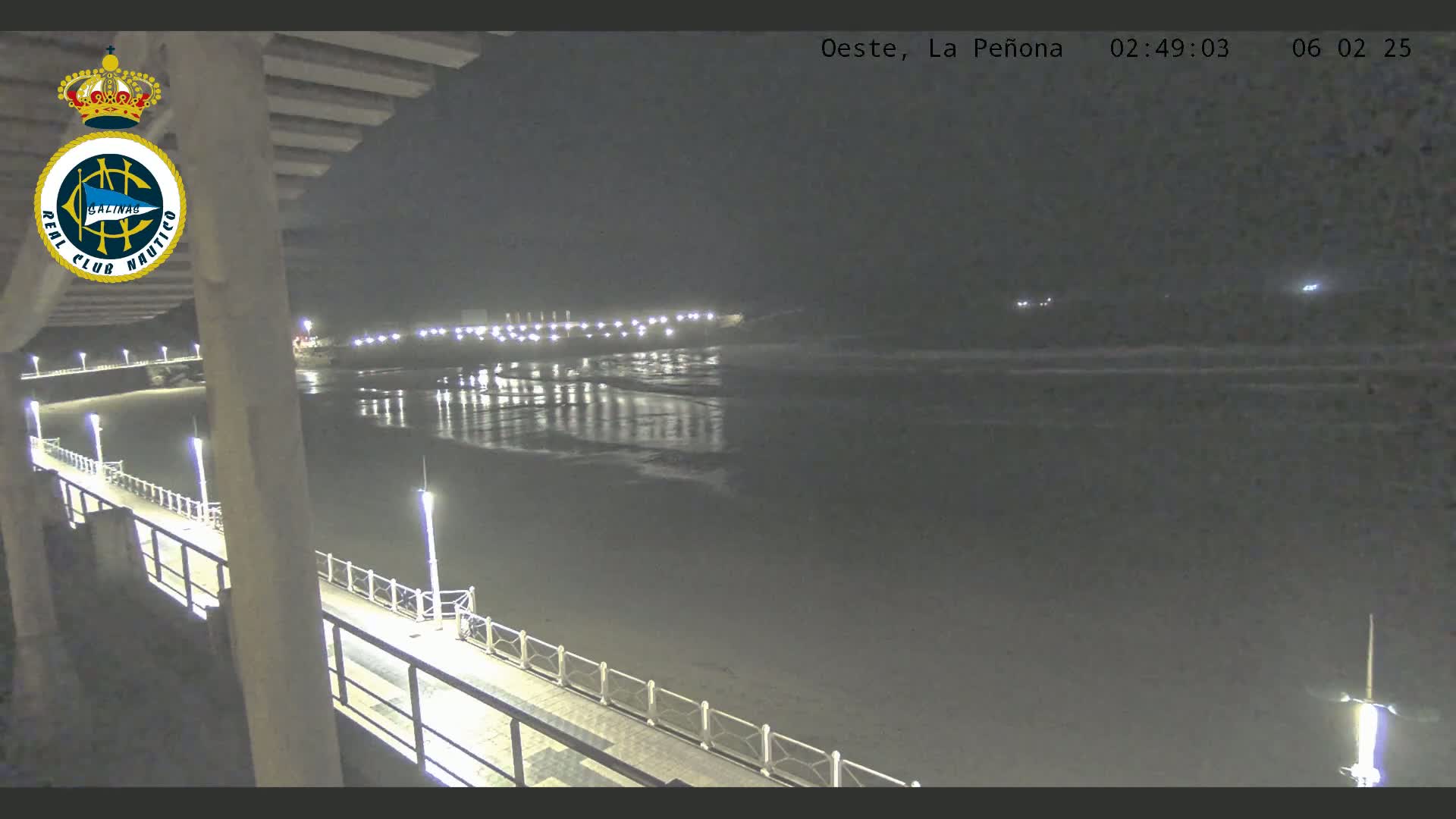 A nighttime view from a pier shows a dark beach and calm ocean water reflecting the lights of a distant shore.