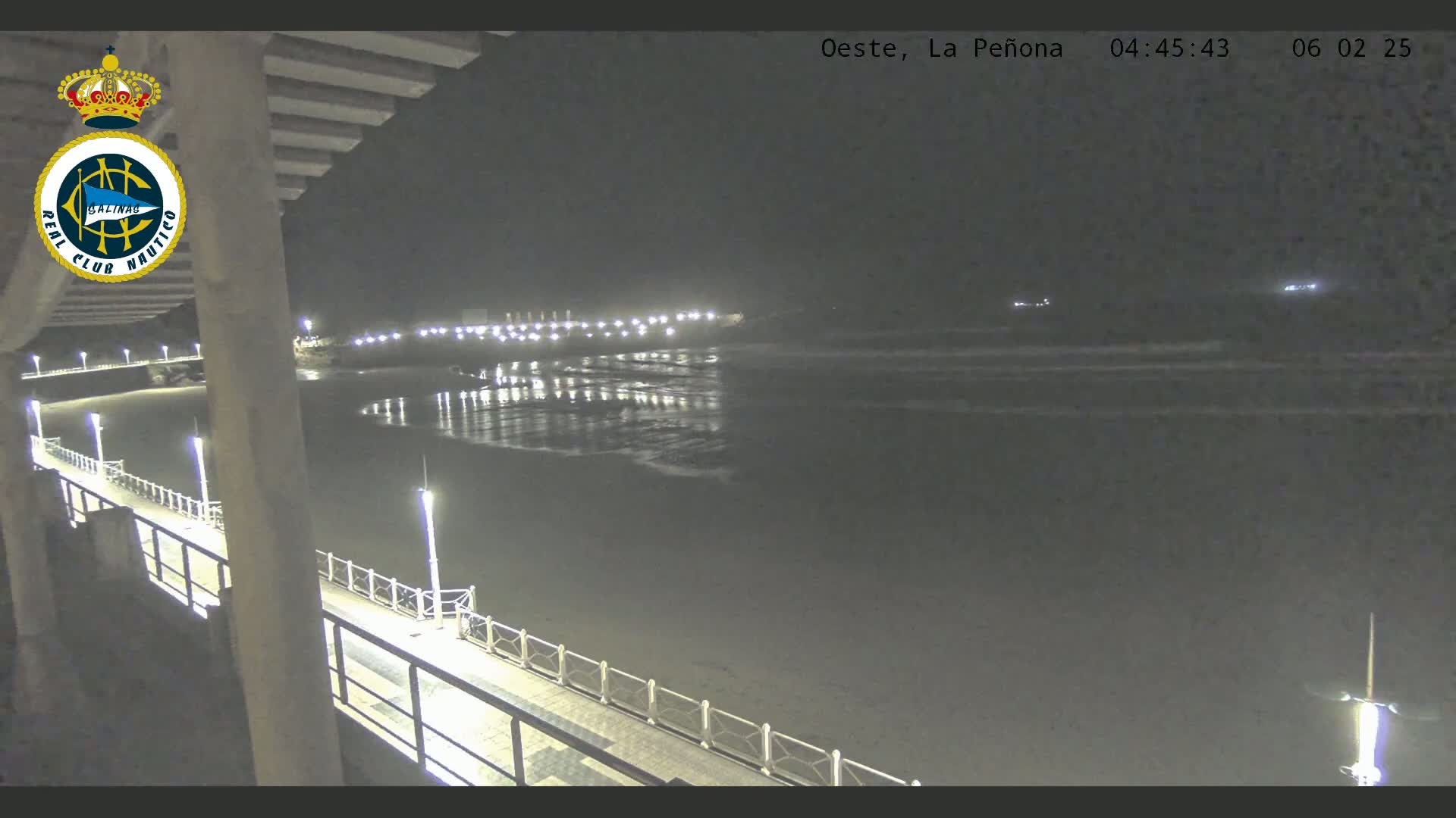 A nighttime view from a pier shows a dark beach and calm ocean with lights reflecting on the water, and lights illuminating a walkway along the pier.