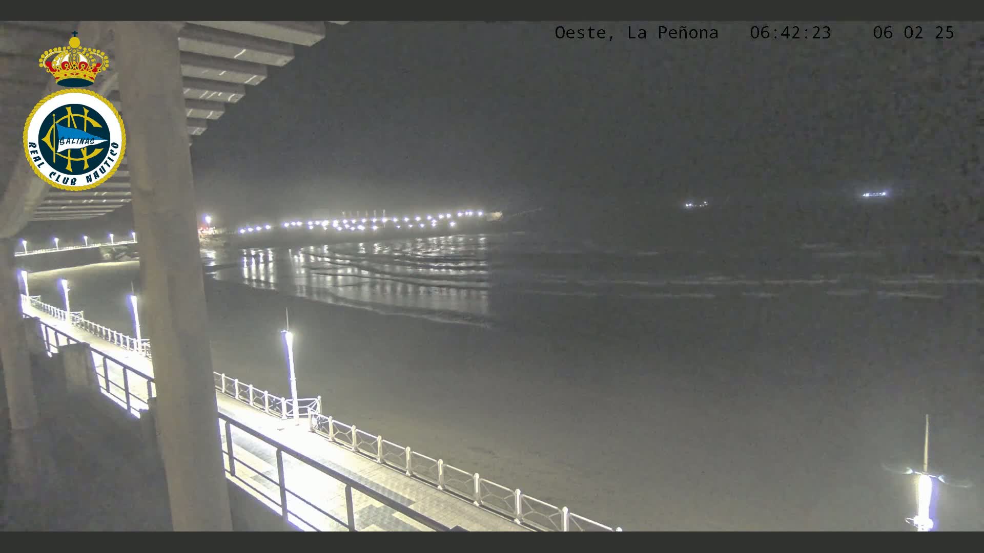 A nighttime view from a pier shows a dark beach with gentle waves lapping the shore, illuminated by lights along the pier and in the distance.