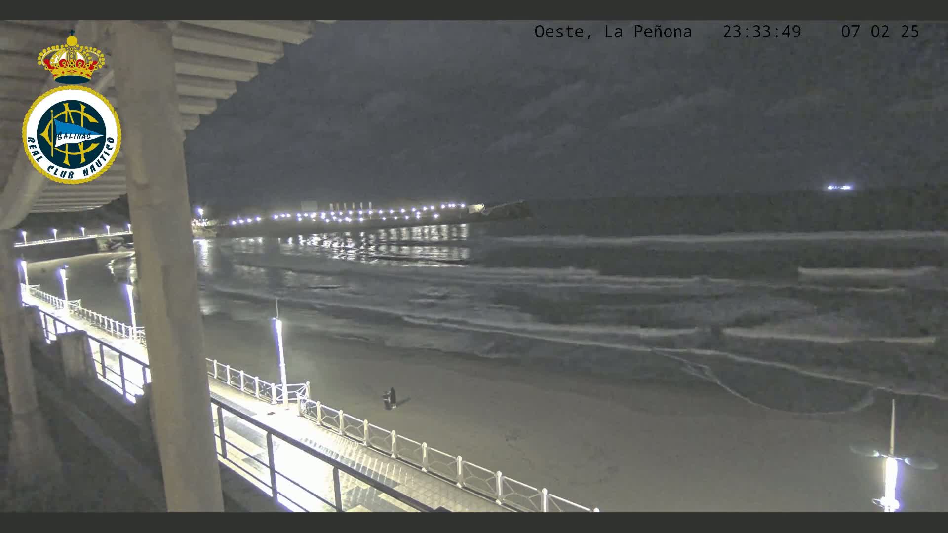 A nighttime view shows a beach with gentle waves lapping the shore under a cloudy sky, with a pier extending from the foreground into the water, illuminated by lights.