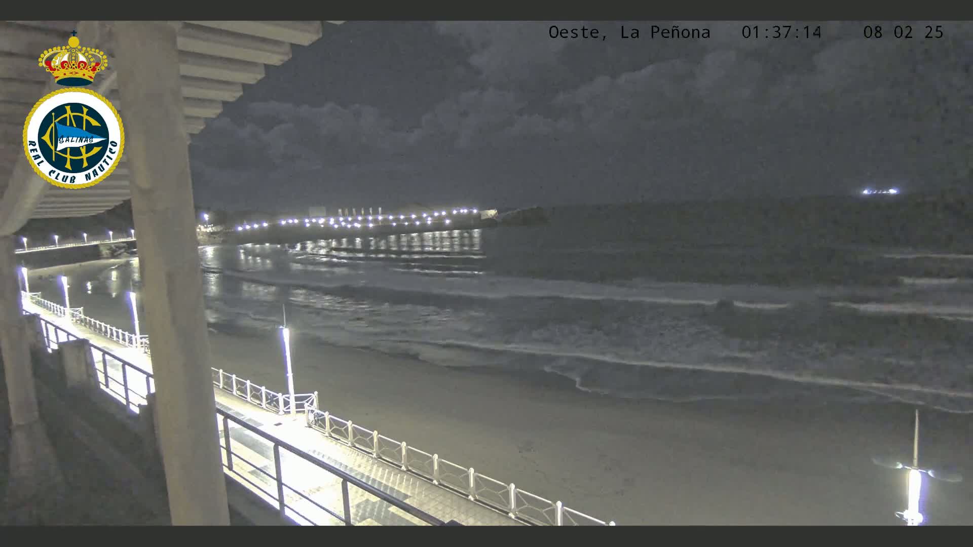 A nighttime view from an elevated position shows a beach with gentle waves lapping the shore under a partly cloudy sky, and a pier extending into the water.