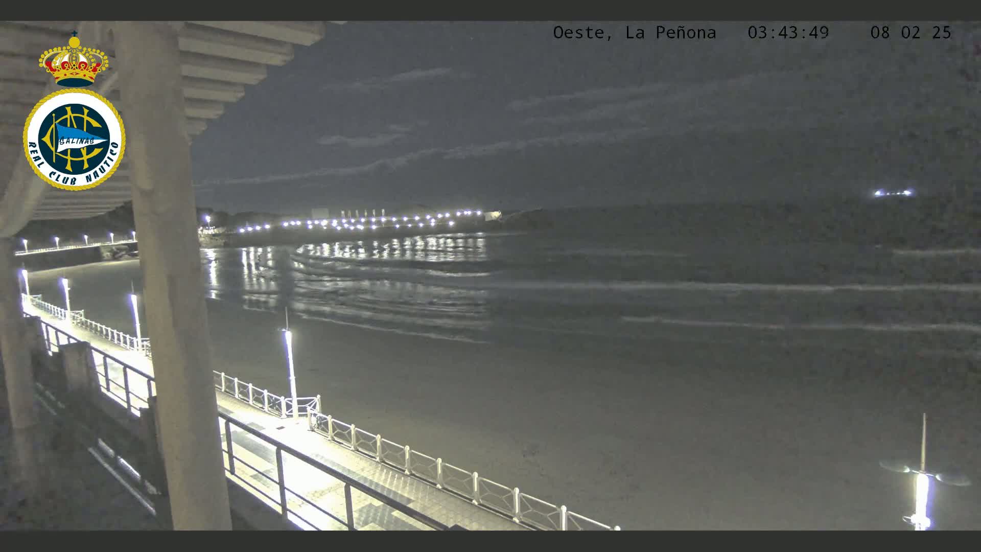A nighttime view from a pier shows gently rolling waves lapping a dark sandy beach under a mostly cloudy sky.