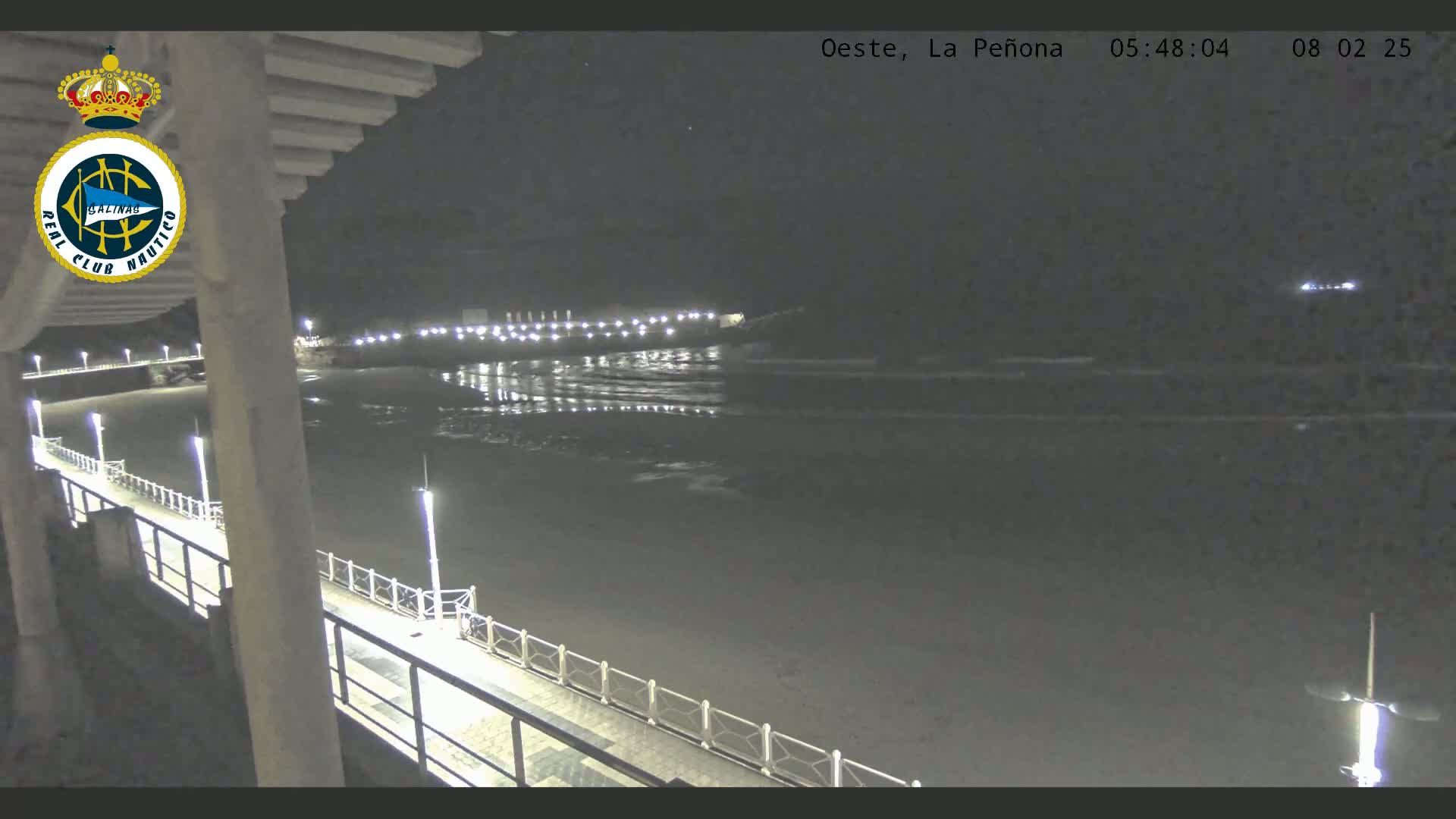 A nighttime view from a pier shows a dark beach and calm ocean under a clear sky, with lights illuminating the pier and the shoreline in the distance.