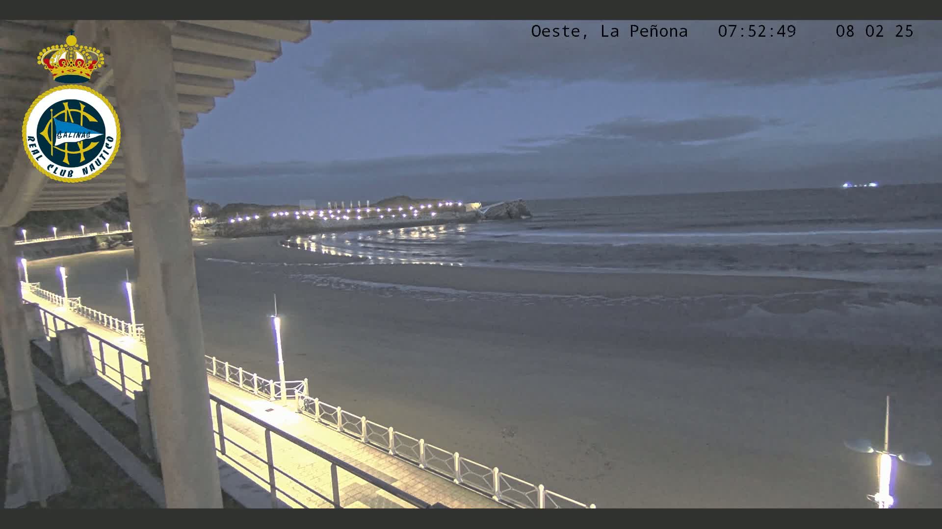 A dimly lit beach at twilight shows a long walkway with lights leading to a rocky outcropping in the water, with gentle waves lapping the shore under a mostly cloudy sky.