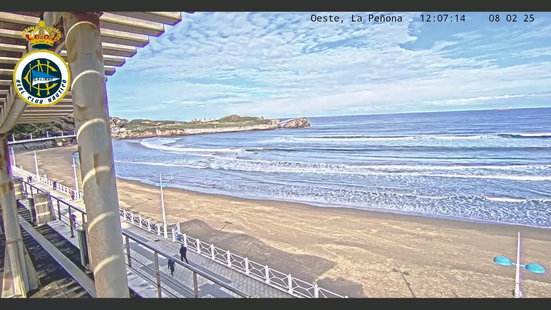 A sandy beach with gentle waves under a partly cloudy sky is viewed from an elevated vantage point, showing a boardwalk and people walking along it.