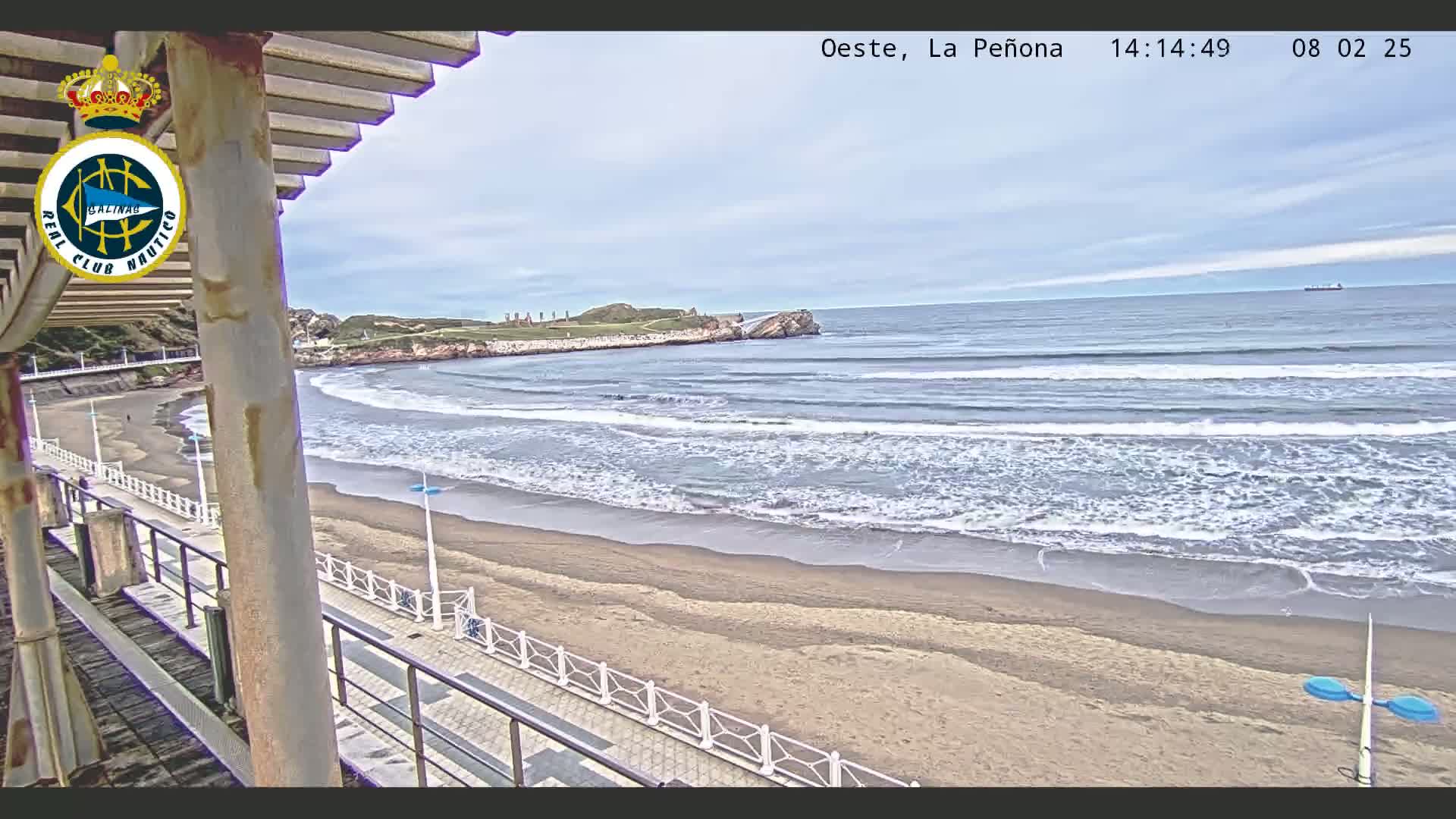 A sandy beach with gentle waves under a mostly cloudy sky is viewed from a boardwalk with a partial view of a building's structure.