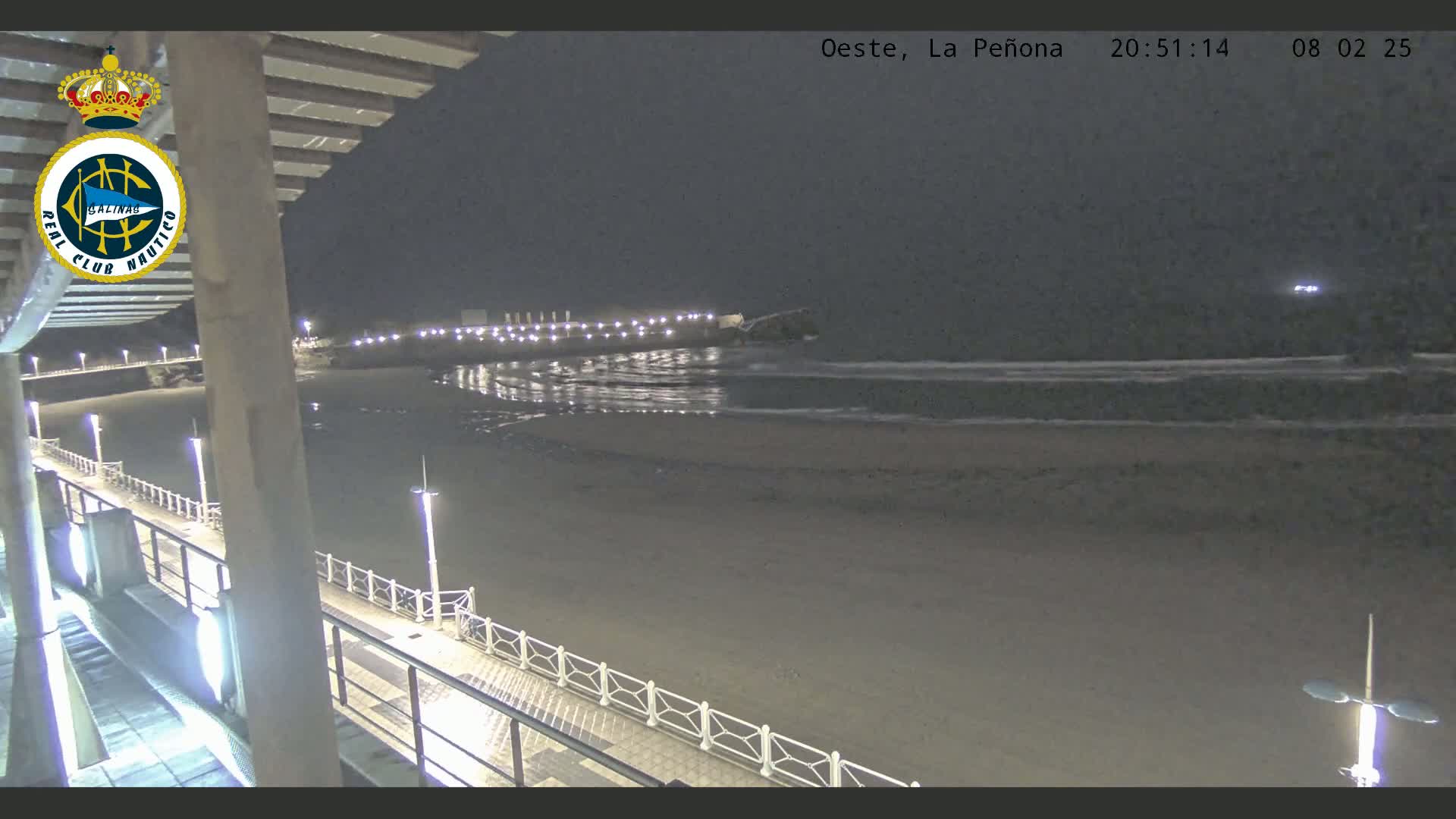 A nighttime view of a beach at low tide, seen from a pier with lights illuminating the walkway and the shore.