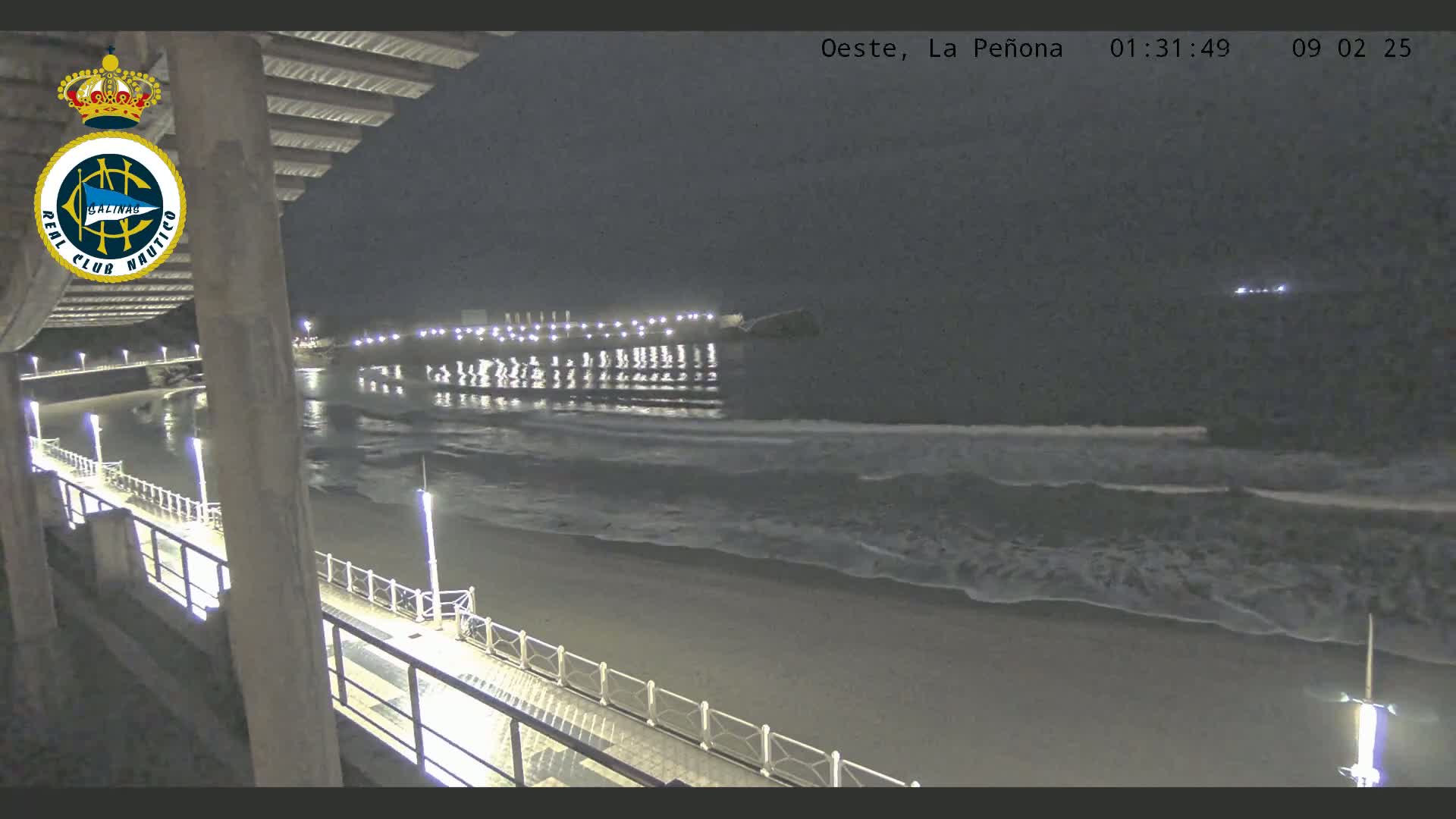 A nighttime view of a beach with gentle waves lapping the shore, a pier extending into the water, and lights illuminating the scene.
