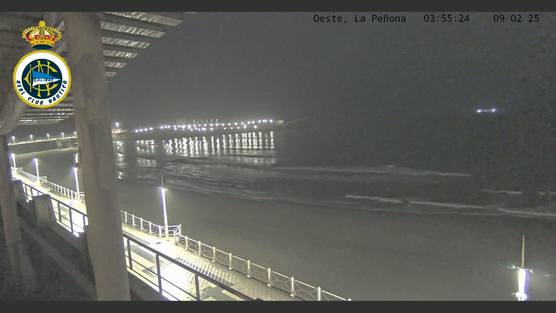 A nighttime view from a pier shows a dark beach and gently lapping waves under a mostly clear sky.
