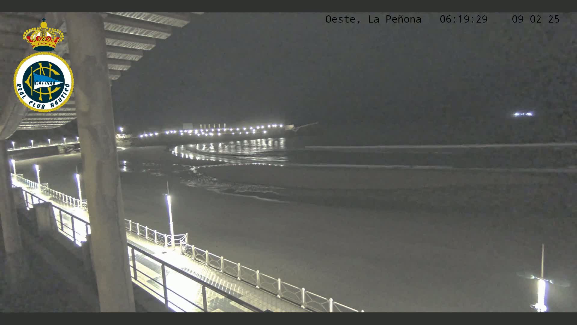 A nighttime view of a beach with gentle waves lapping the shore, seen from a pier with lights illuminating the walkway.