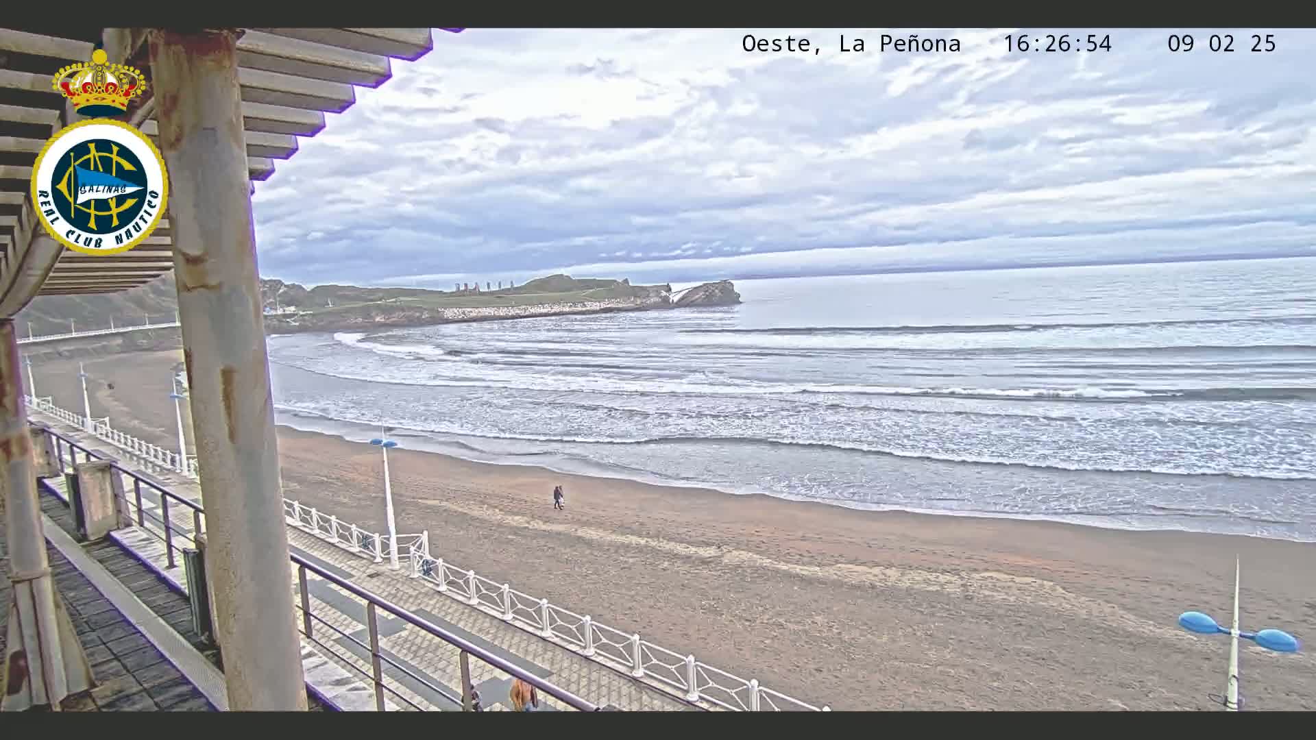 A sandy beach with gentle waves under a cloudy sky is viewed from a seaside structure.