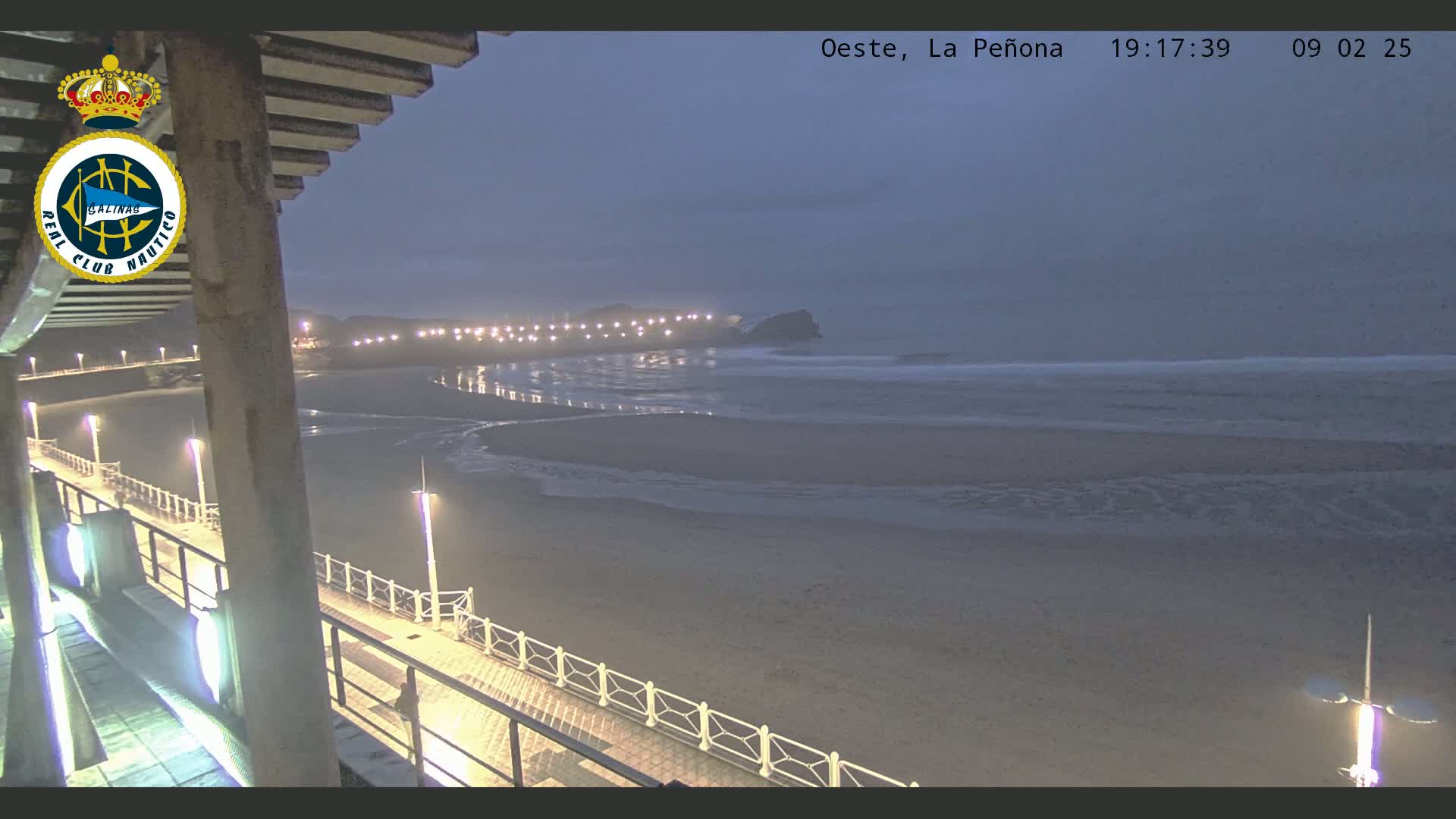 A nighttime view of a beach with gentle waves under a dusky sky, seen from a pier with lights illuminating the walkway.
