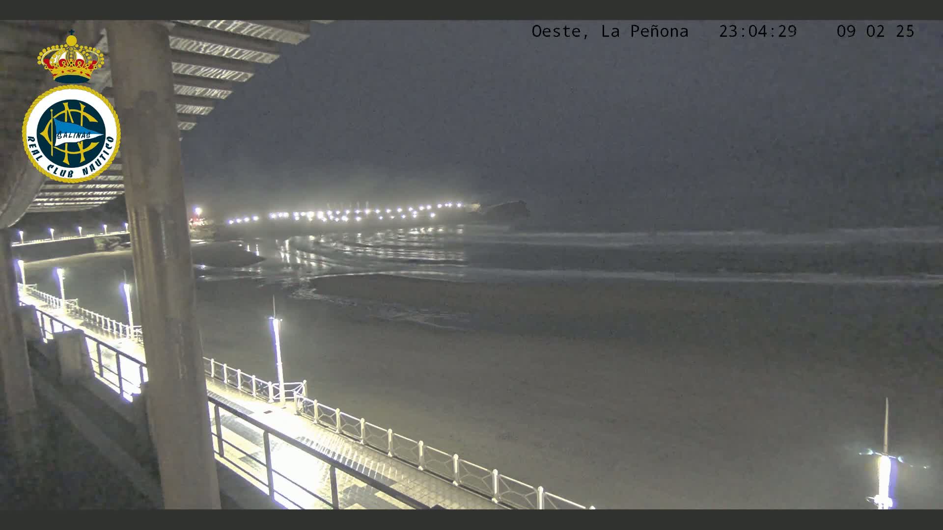 A nighttime view from a pier shows a dark, sandy beach with gentle waves lapping the shore under a hazy sky.