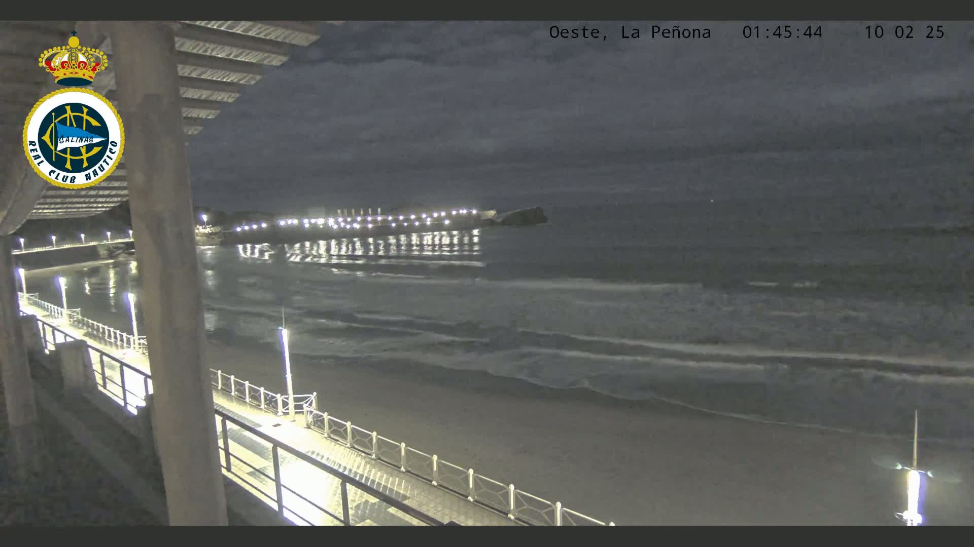 A nighttime view from a pier shows a dark, sandy beach with gentle waves lapping the shore under a cloudy sky.