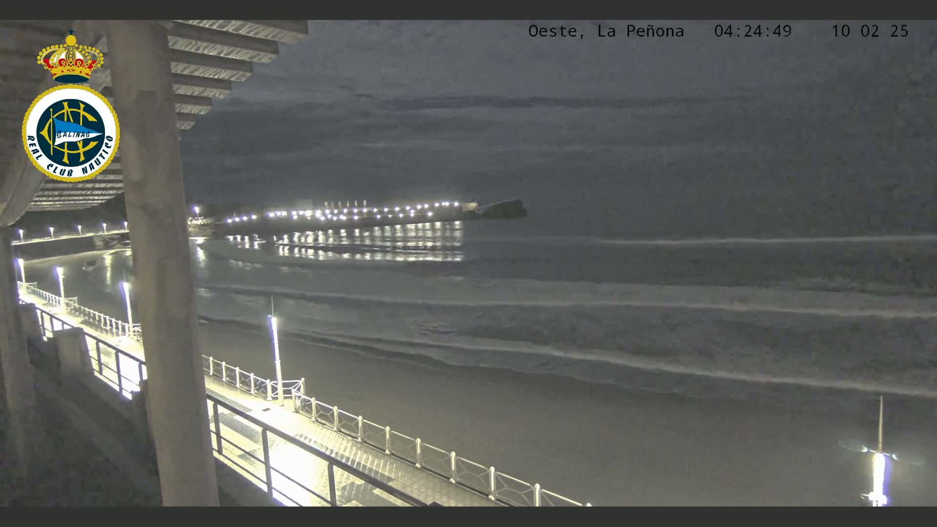 A nighttime view from a pier shows a calm ocean with gentle waves lapping the shore under a dark, cloudy sky.