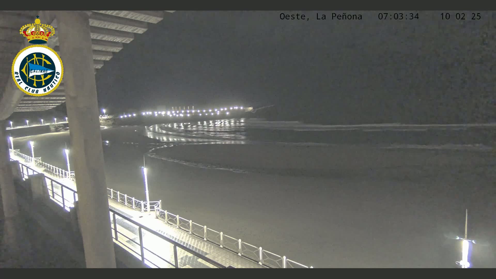 A nighttime view from a pier shows a dark sandy beach with gently lapping waves under a clear, dark sky.