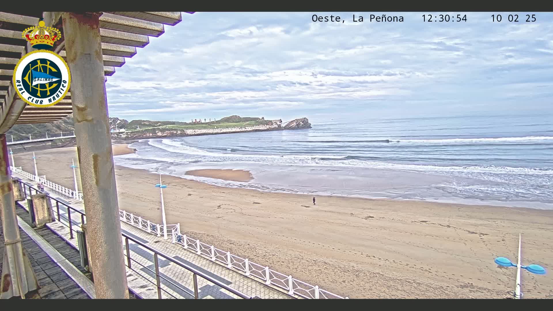 A sandy beach with gentle waves under a partly cloudy sky is viewed from an elevated vantage point, showing a boardwalk and a rocky headland in the distance.