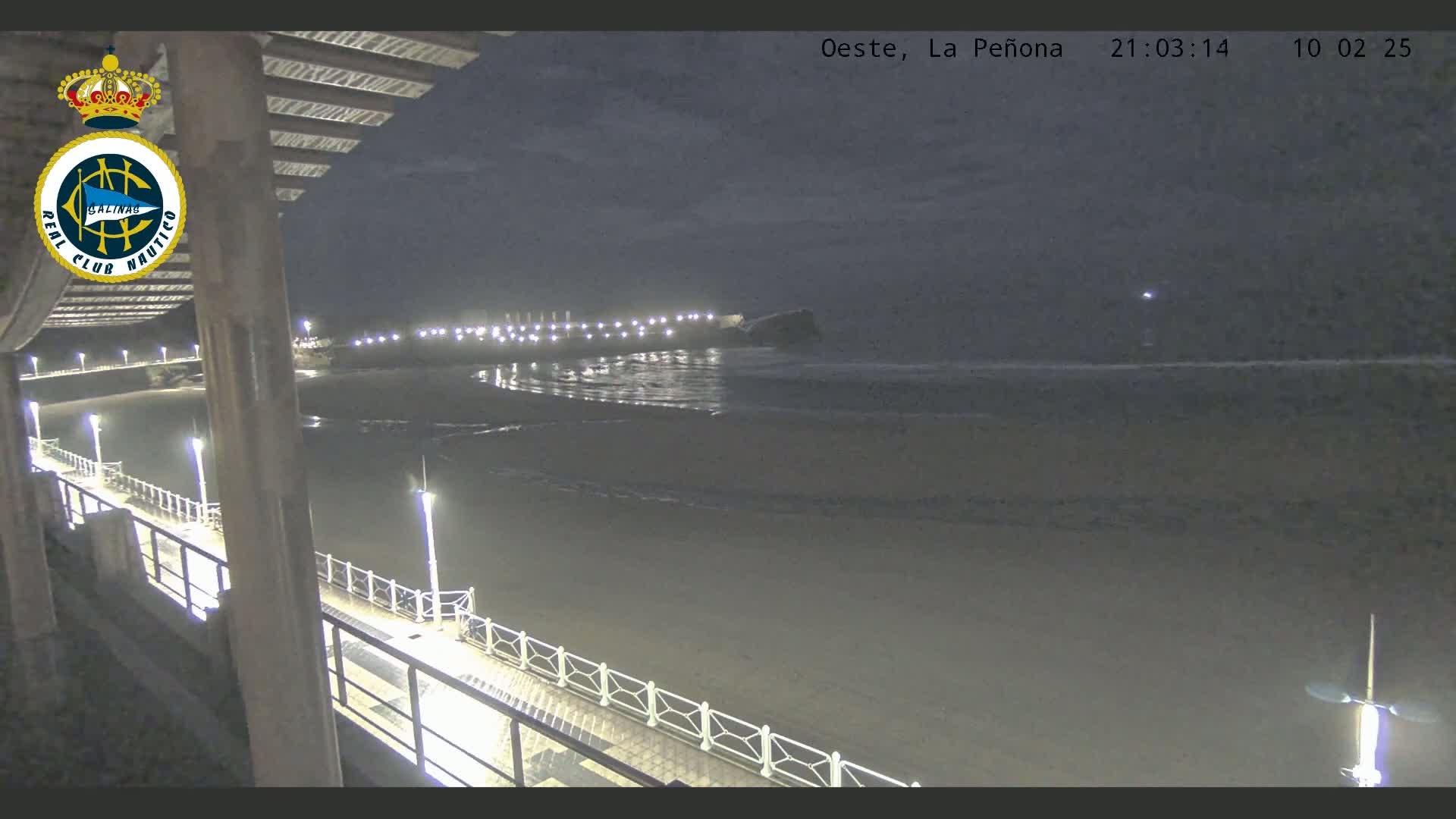 A nighttime view from a pier shows a dark, sandy beach with gentle waves lapping the shore under a mostly clear sky.