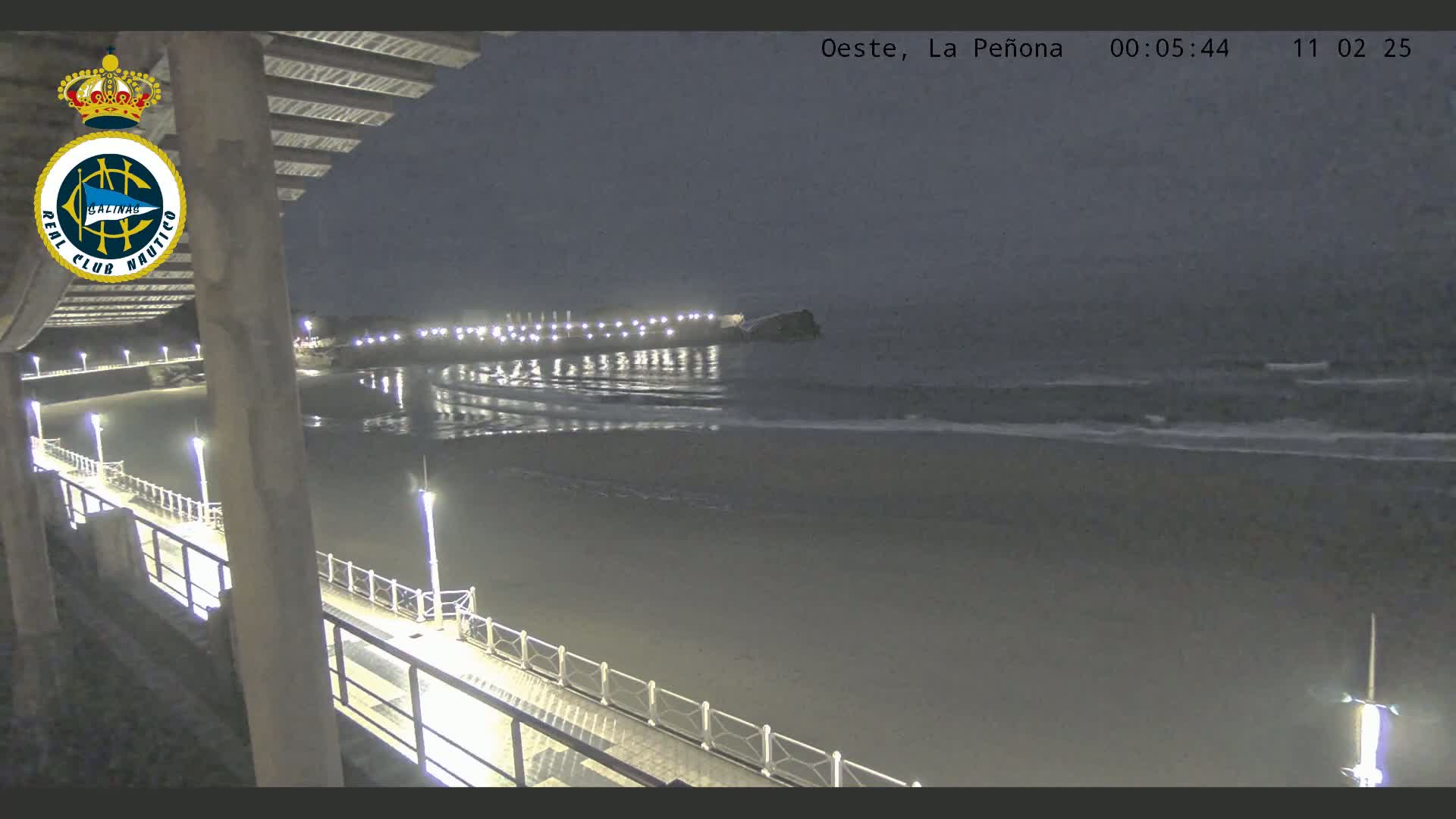 A nighttime view from a pier shows a calm ocean with gentle waves lapping at a dimly lit beach.