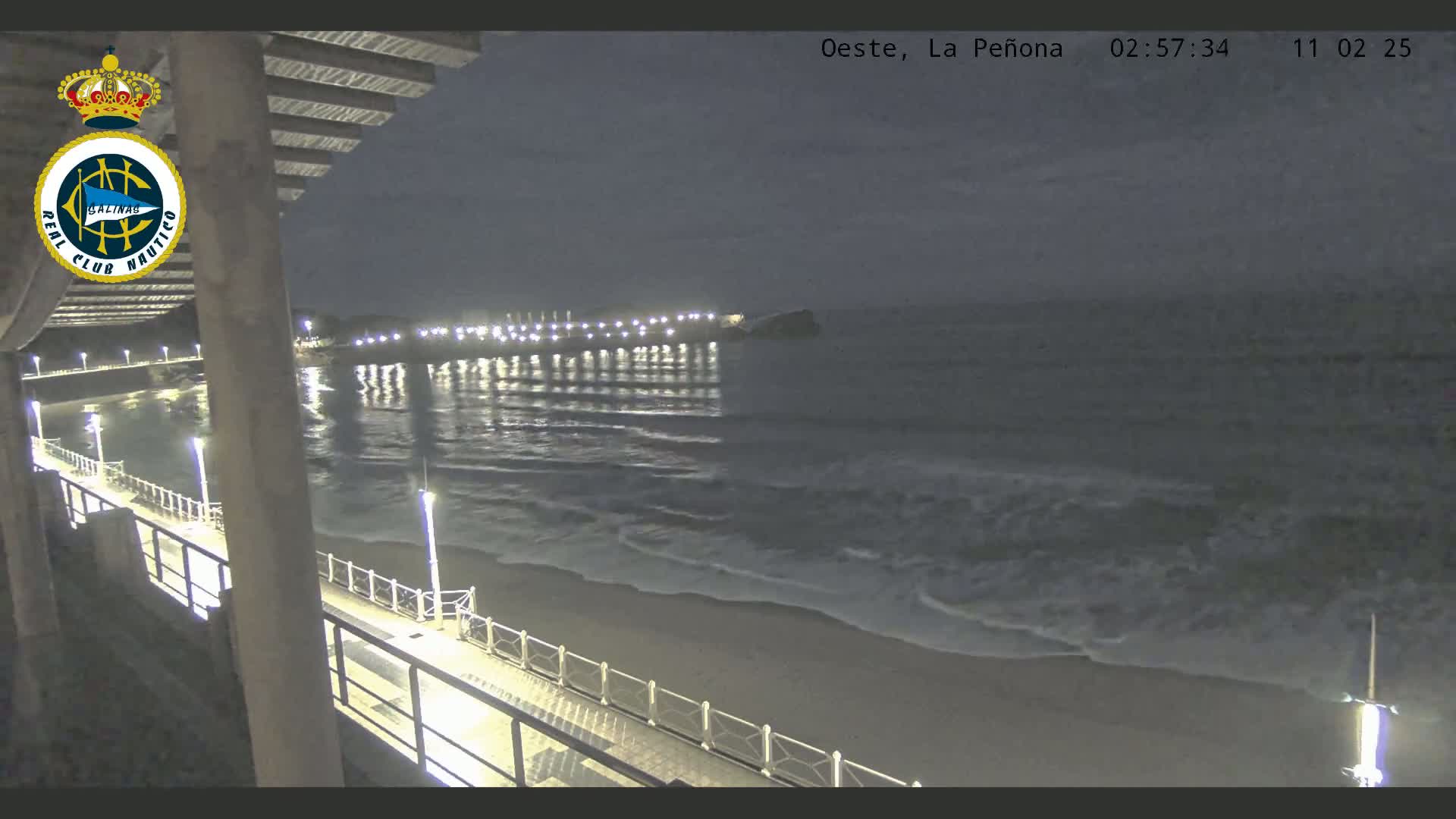 A nighttime view from a pier shows a gently lapping ocean under a dark, cloudy sky.
