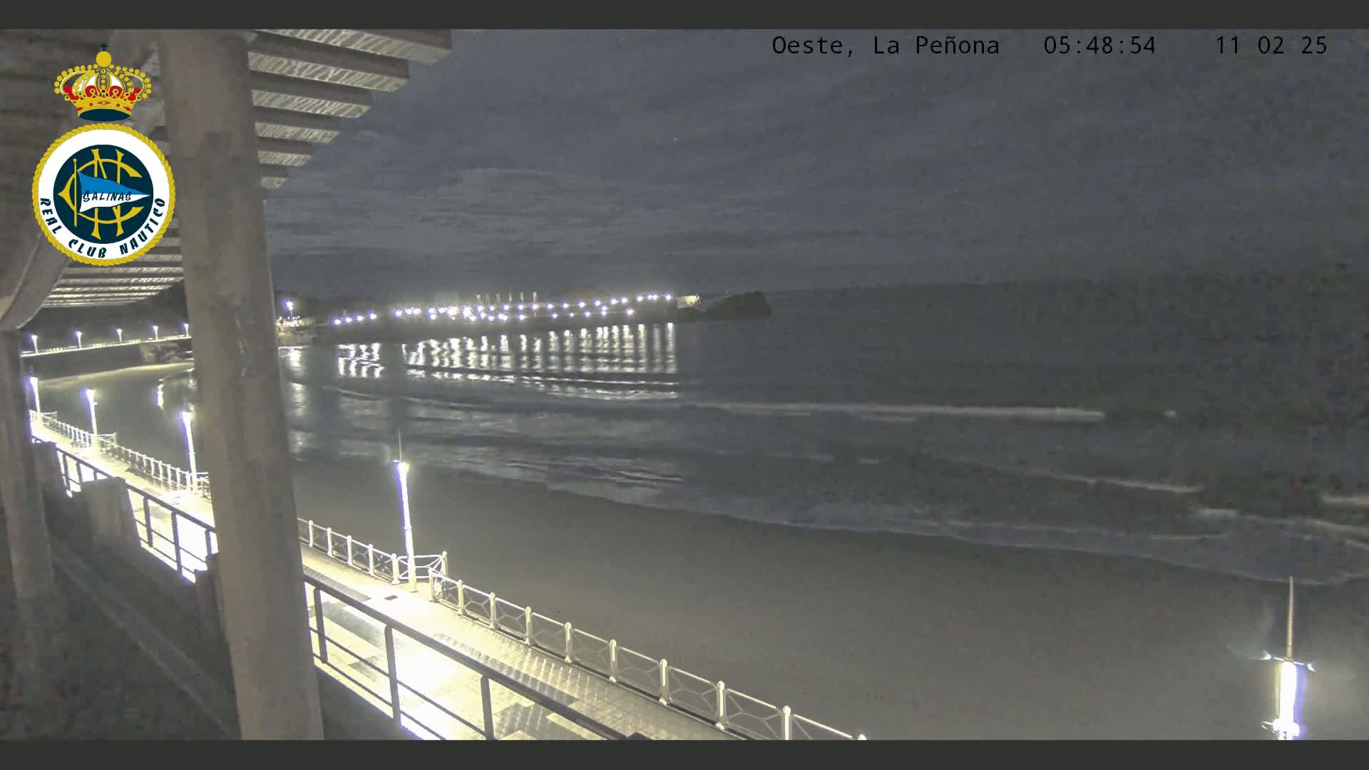 A nighttime view from a pier shows a beach with gentle waves under a dark sky, and lights illuminating the pier and a distant shoreline.