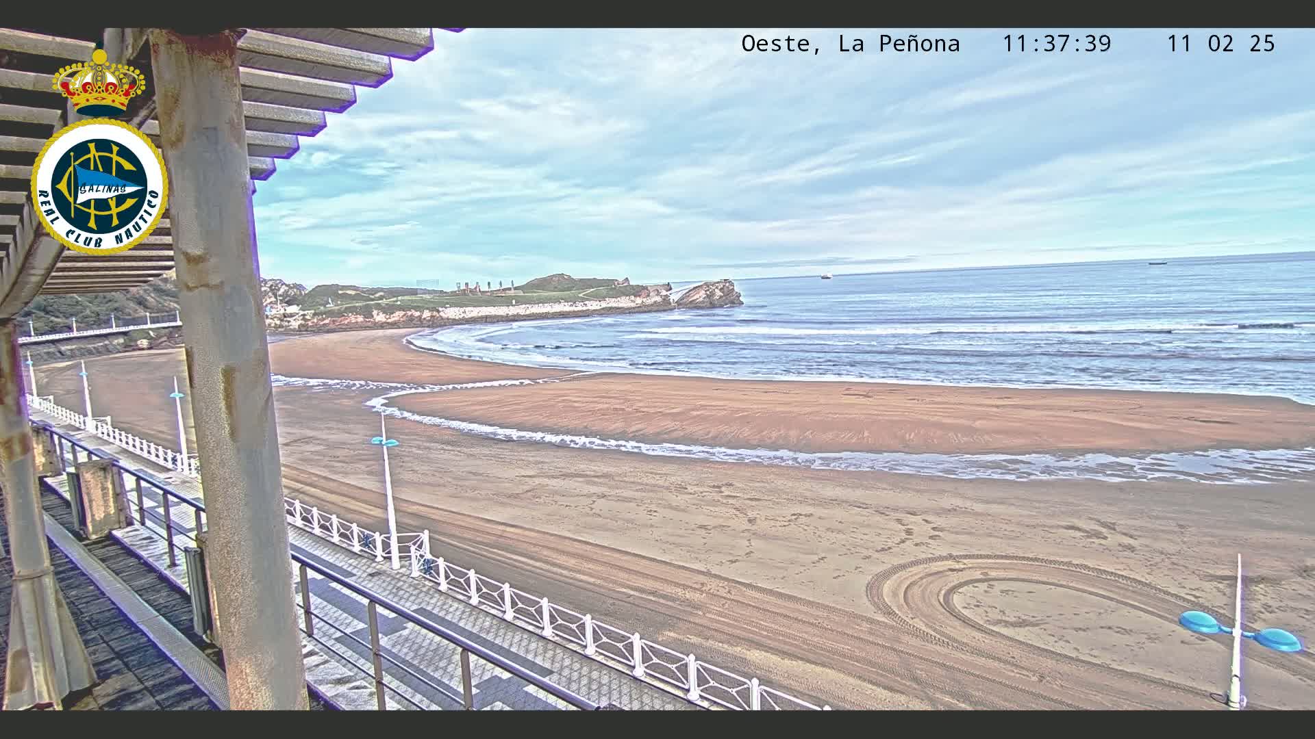 A sandy beach with gentle waves under a partly cloudy sky is viewed from an elevated vantage point, showing a boardwalk and a portion of a structure with a pergola.
