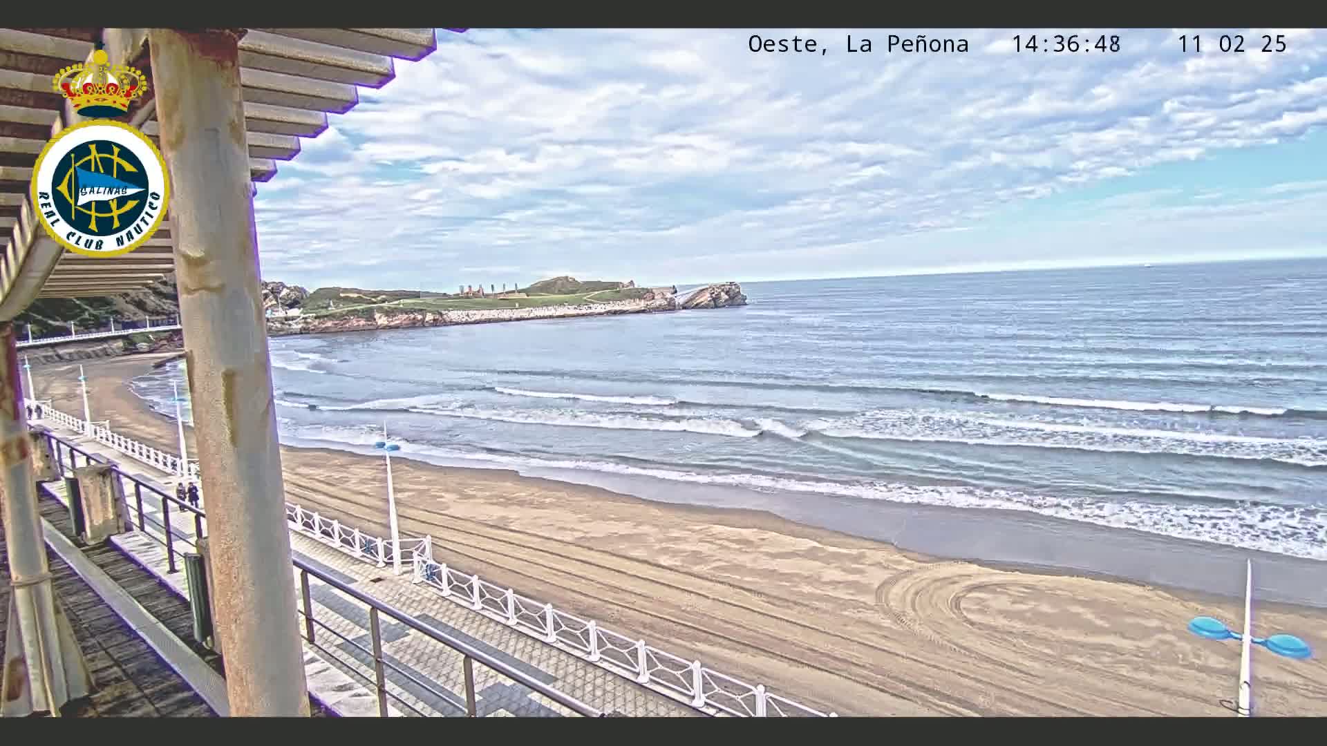 A partly cloudy day shows a sandy beach with gentle waves lapping the shore, viewed from a seaside structure.