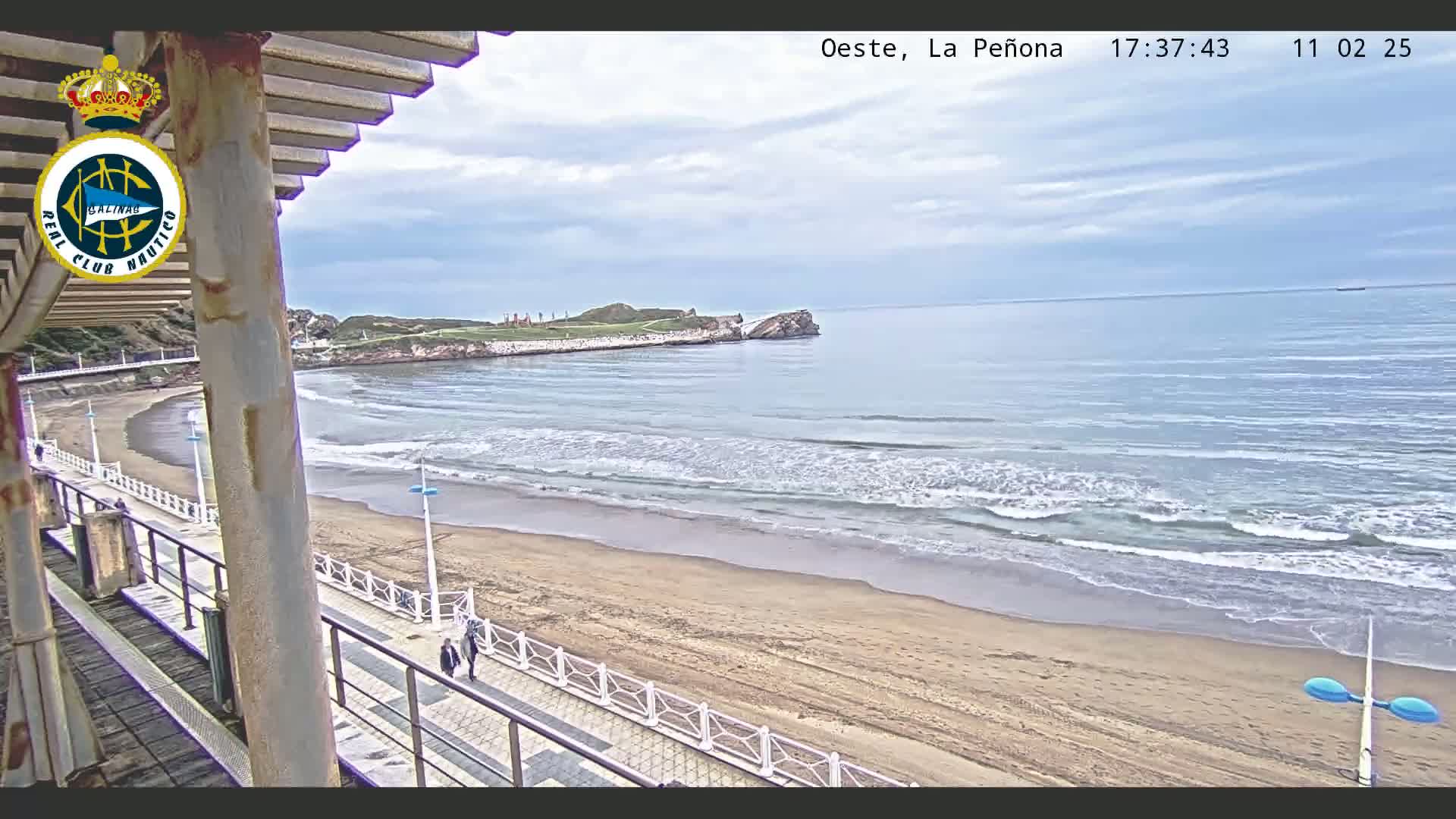 A sandy beach with gentle waves stretches out to a calm ocean under a mostly cloudy sky, viewed from an elevated position near a boardwalk.