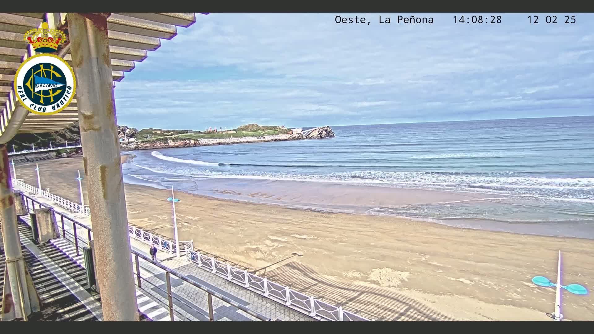 A sandy beach with gentle waves under a partly cloudy sky is viewed from an elevated boardwalk with a railing.