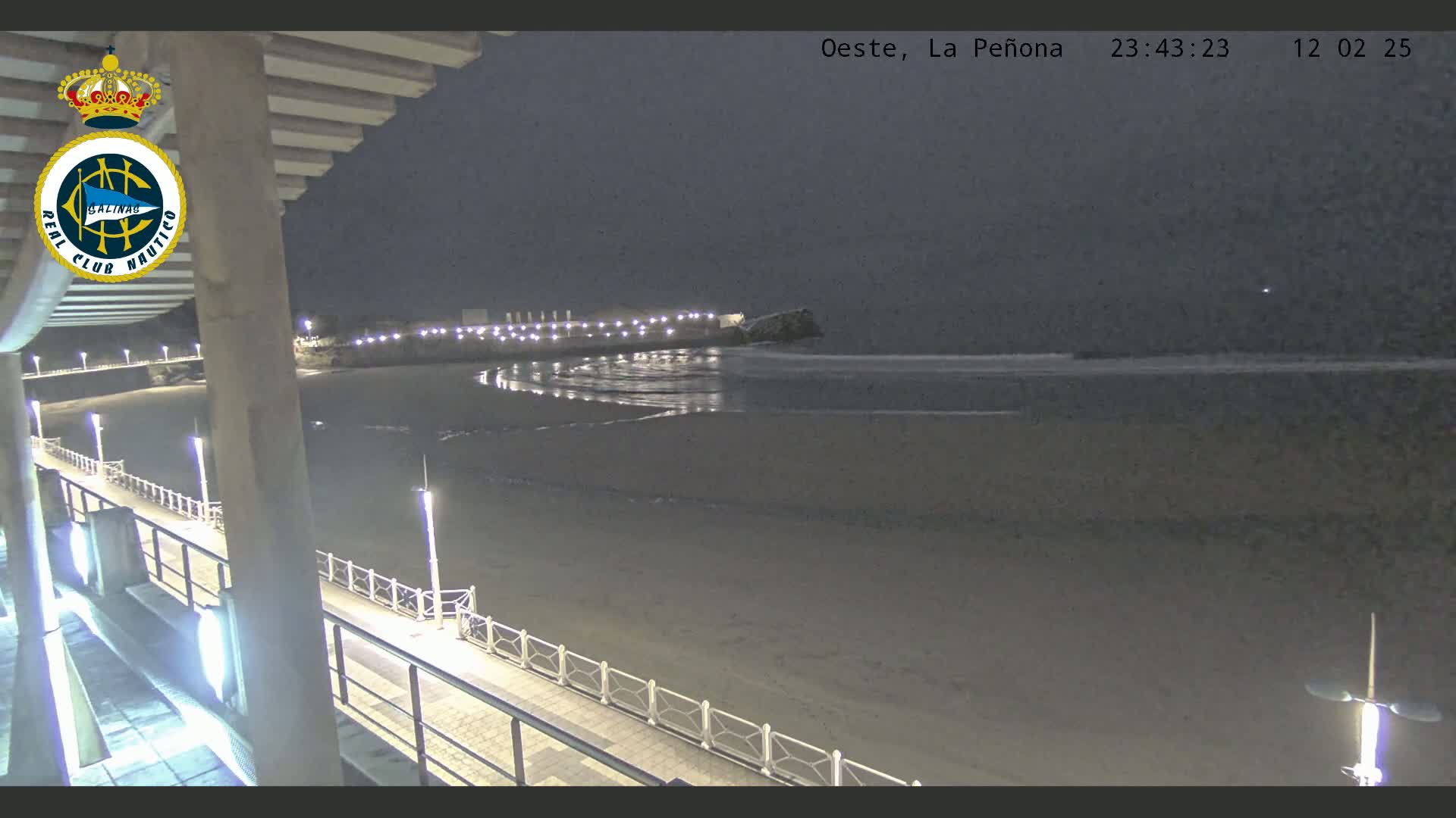 A nighttime view of a beach with gentle waves, illuminated by lights along a walkway and a building in the distance, under a dark sky.