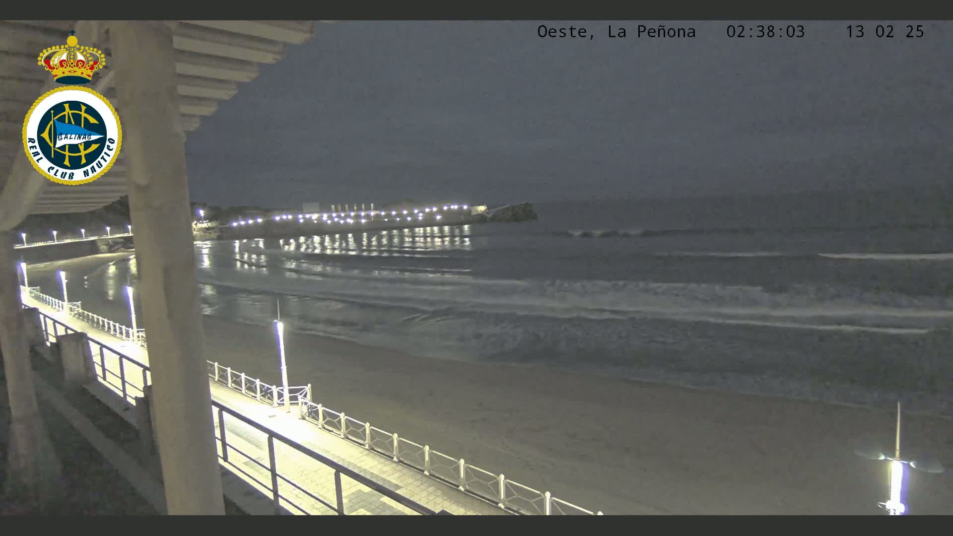 A nighttime view from an elevated position shows a sandy beach, gently lapping waves, and a lit walkway along the shore.