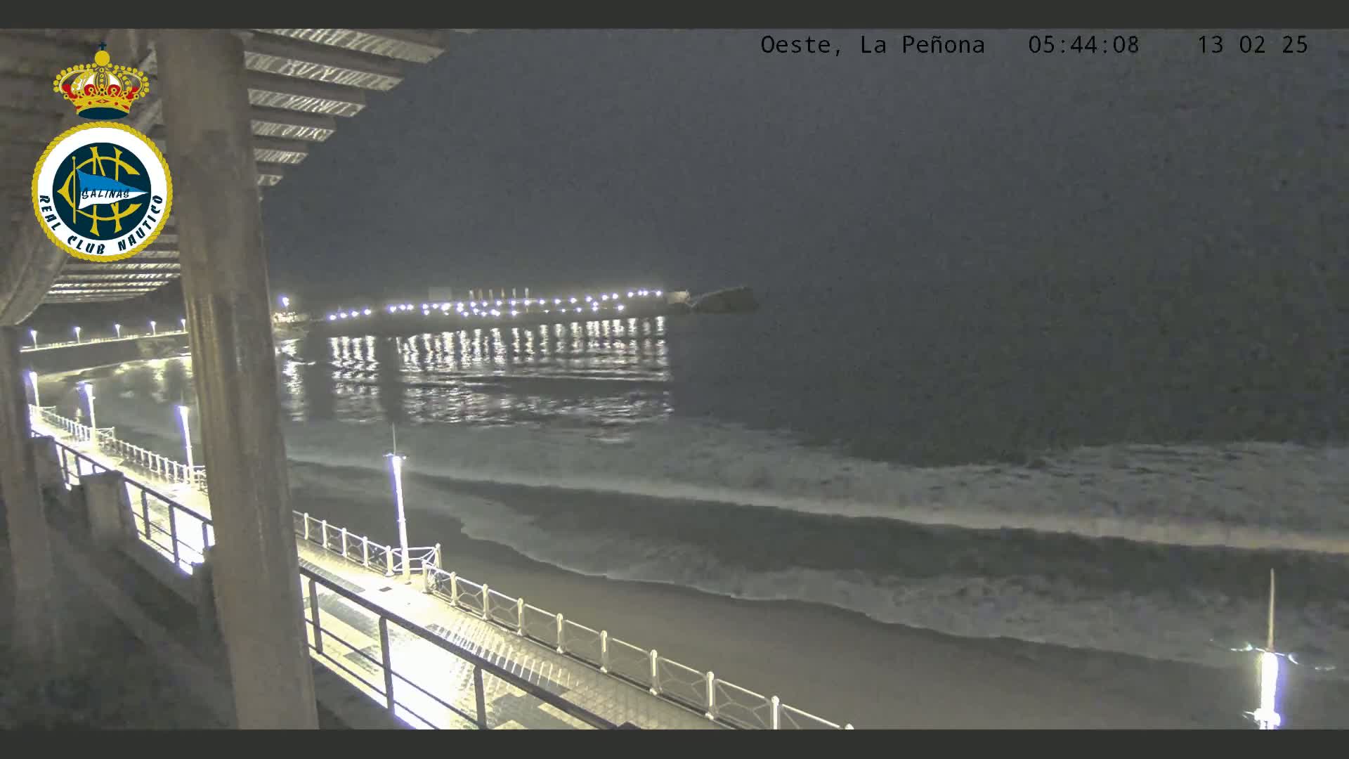 A nighttime view of a beach with gentle waves rolling onto the shore, illuminated by lights along a pier and boardwalk.