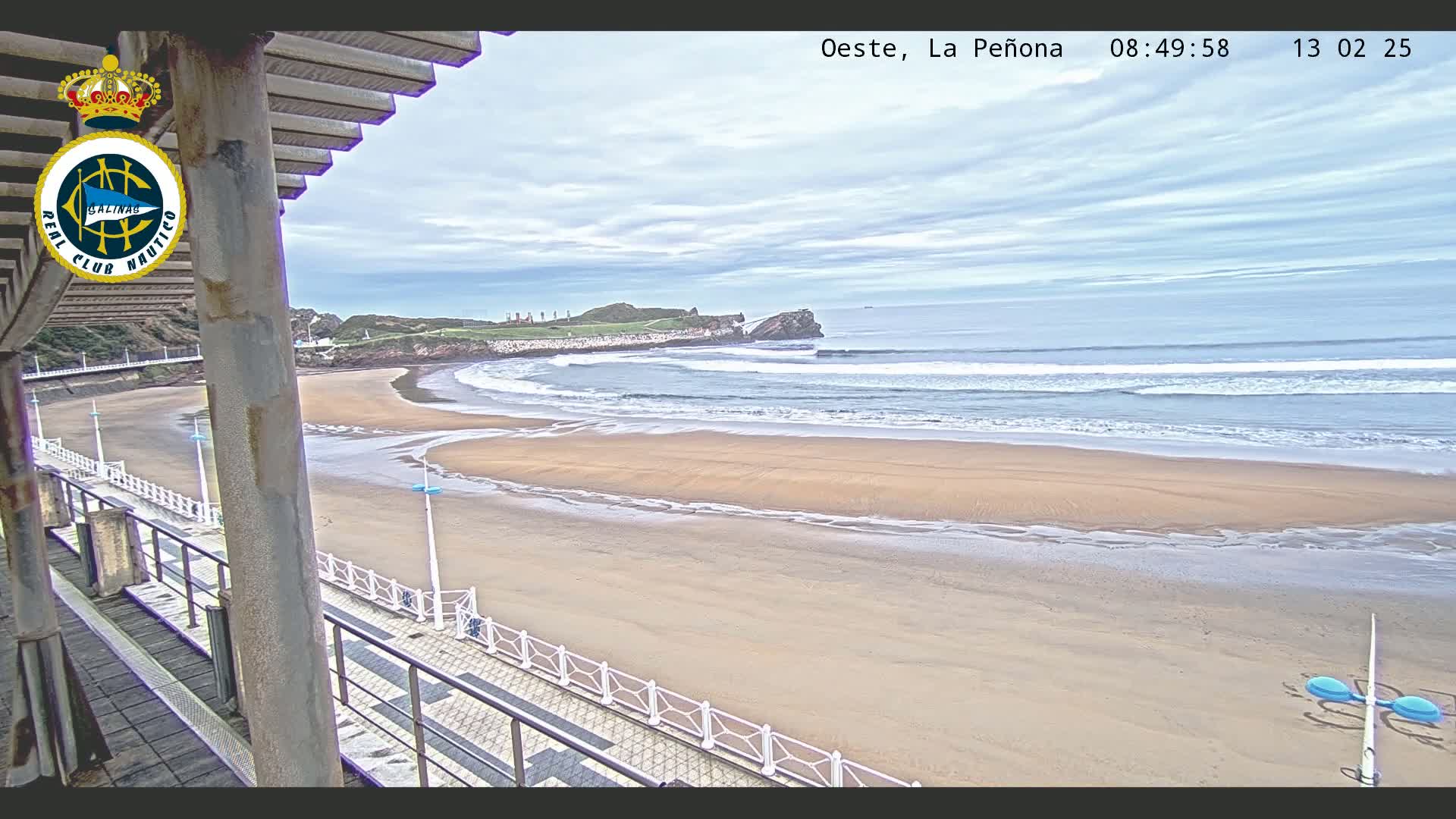 A sandy beach with gentle waves under a cloudy sky is viewed from an elevated position, showing a boardwalk and part of a building in the foreground.