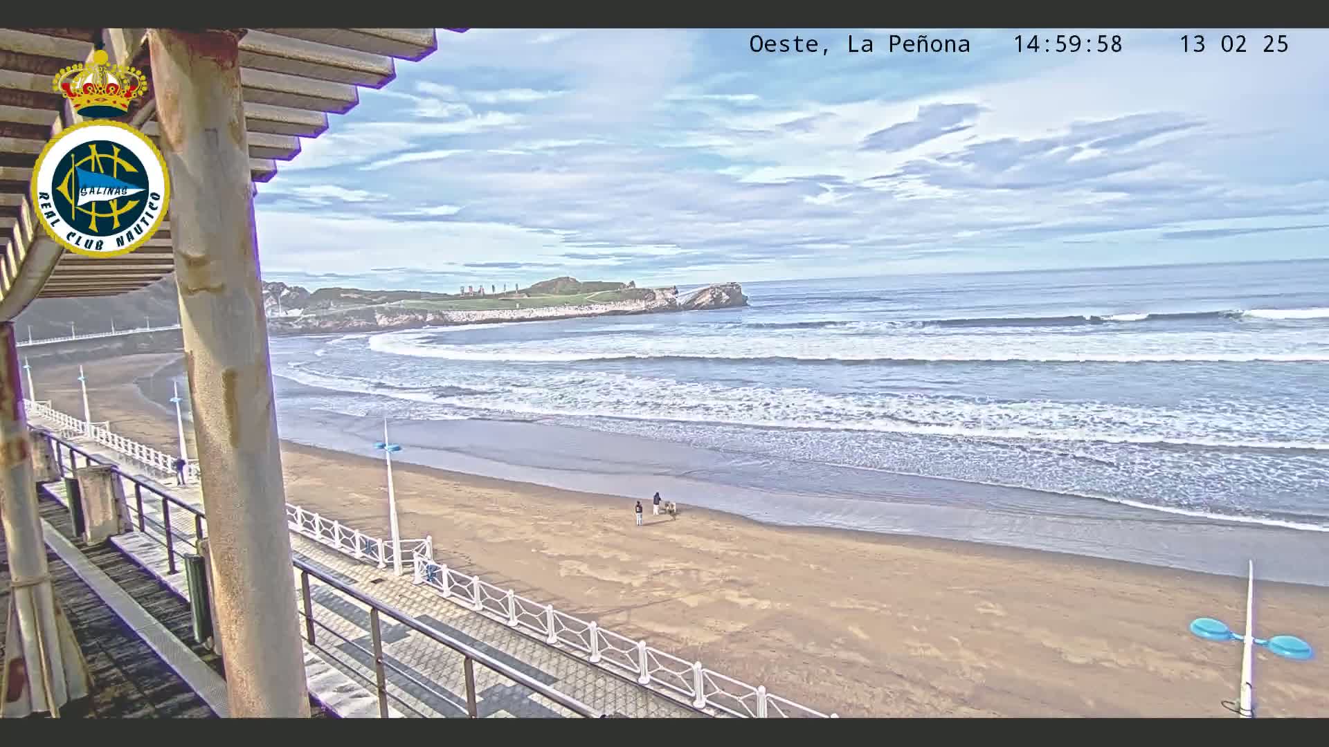 A sandy beach with gentle waves under a partly cloudy sky is viewed from a raised walkway, with a few people and dogs walking near the water's edge.